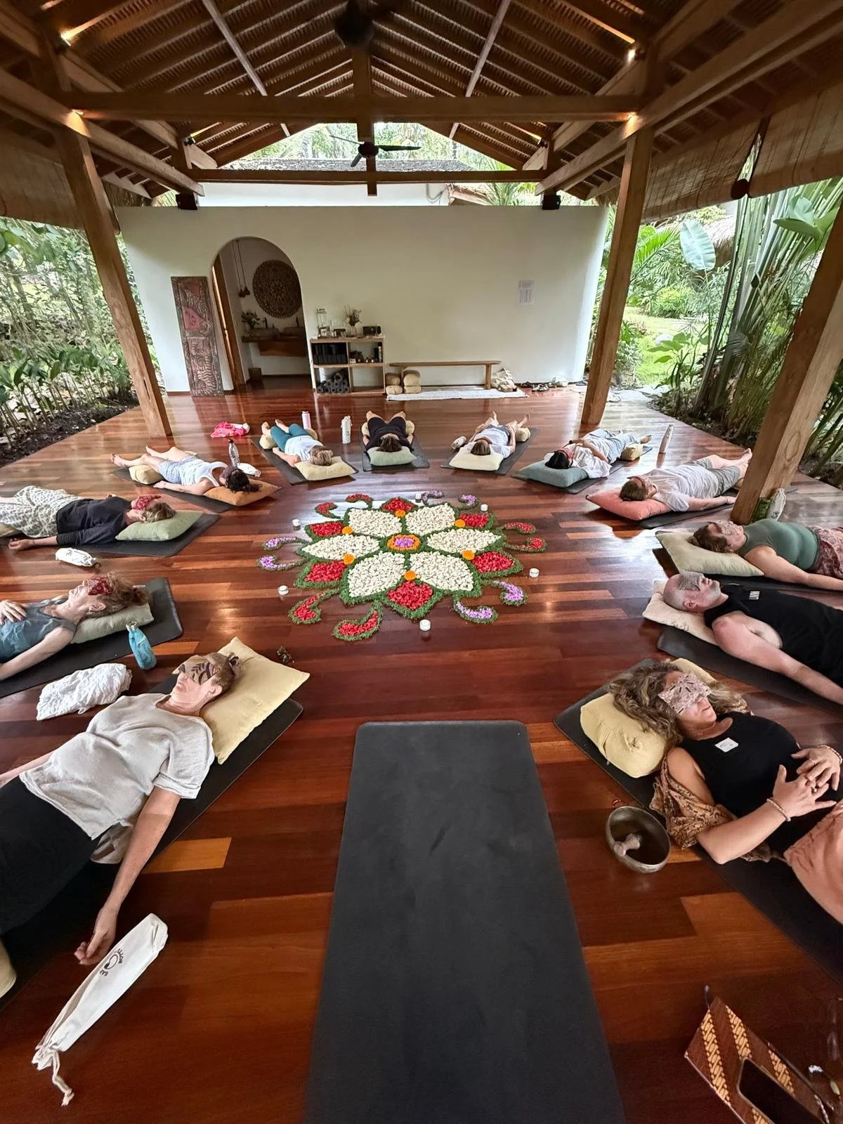 People participating in a group meditation session indoors on wooden floors, arranged in a circle around a floral design, with lush greenery visible outside.