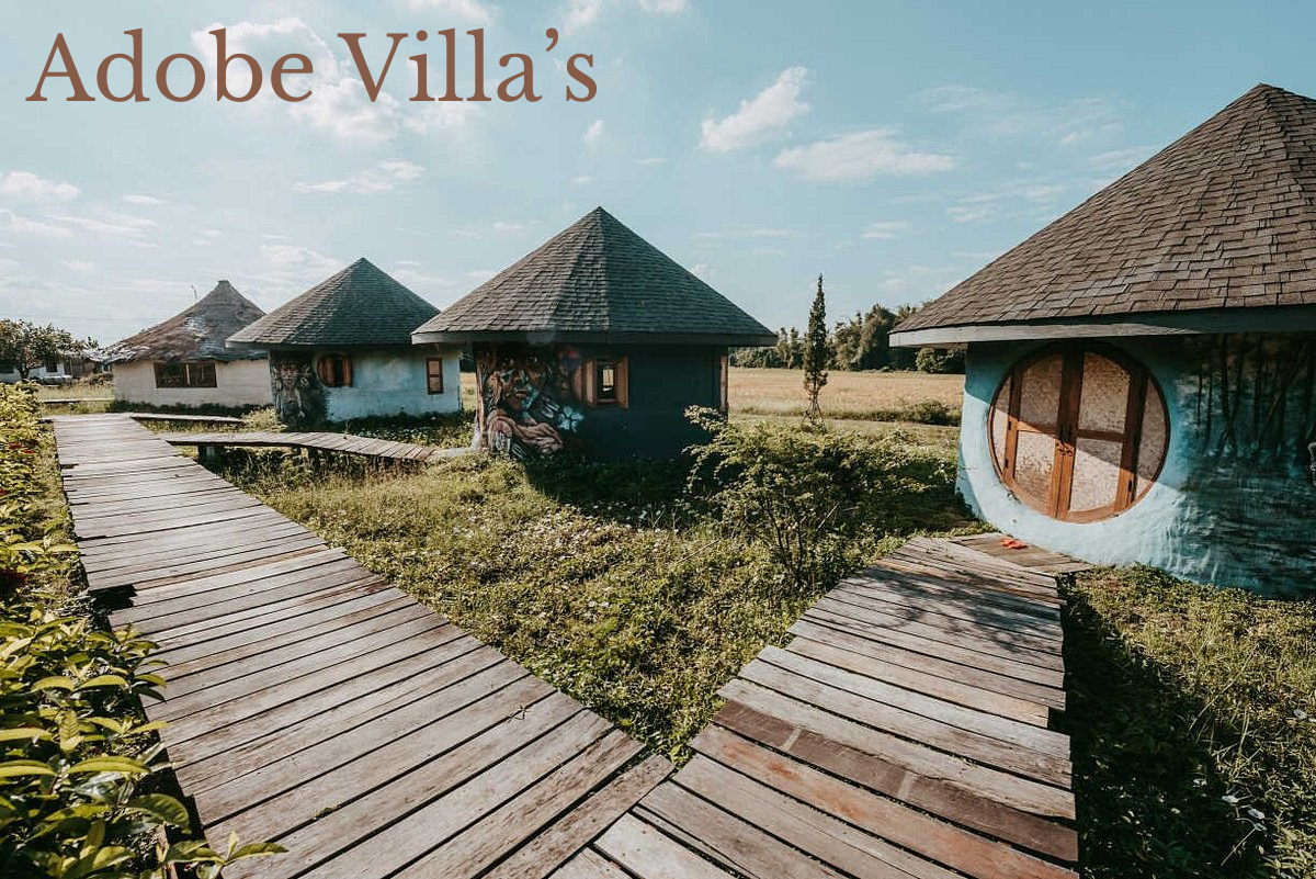 A scenic view of traditional round huts with conical thatched roofs, three of which are visible, set in a grassy landscape with a clear blue sky. A wooden pathway leads through the grass in front of the huts.