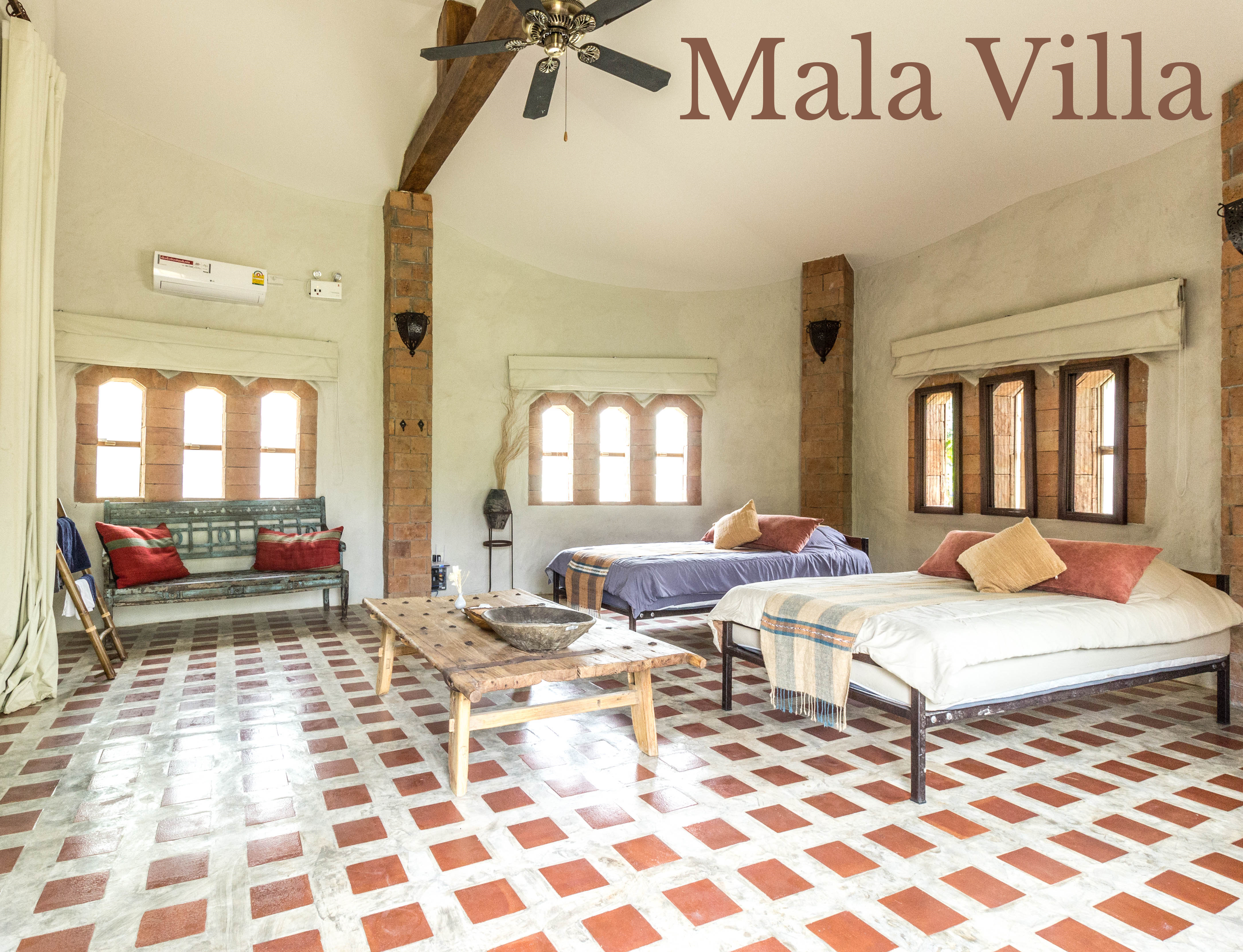 Bright living room with exposed brick columns, white walls, and large windows with white curtains. Contains a wooden coffee table, a metal bed with pillows and throws, a vintage bench, and decorative lamps. A ceiling fan hangs from the ceiling.