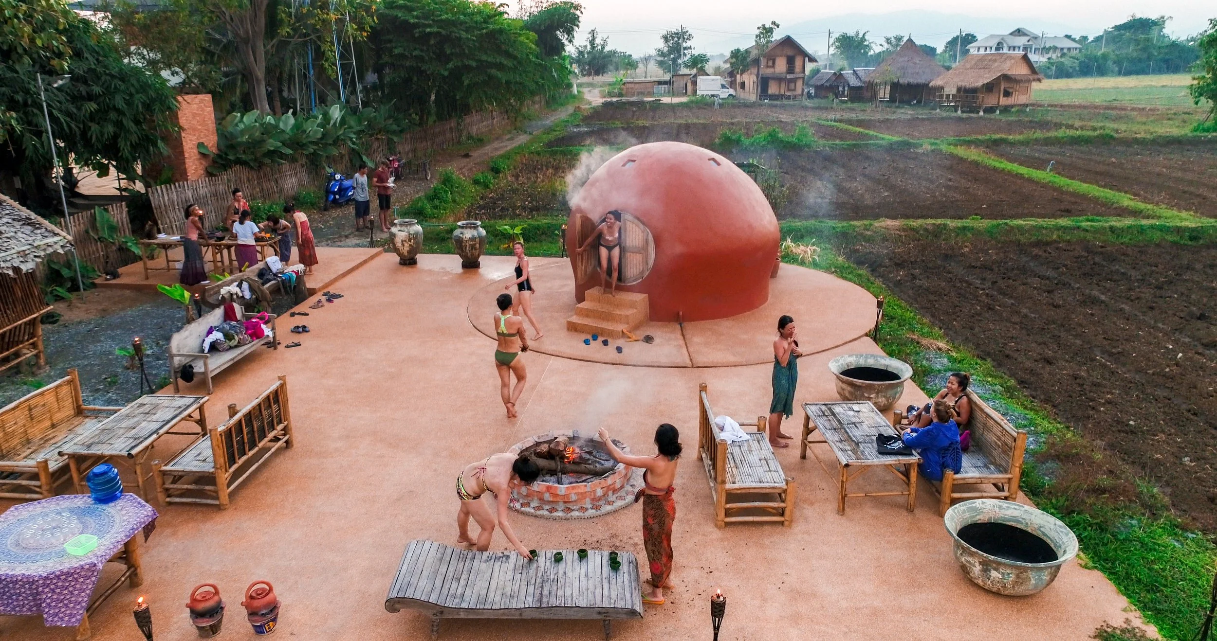 People relaxing and enjoying activities outdoors in a rural setting with a round traditional oven or sauna, a fire pit, and lush green fields in the background
