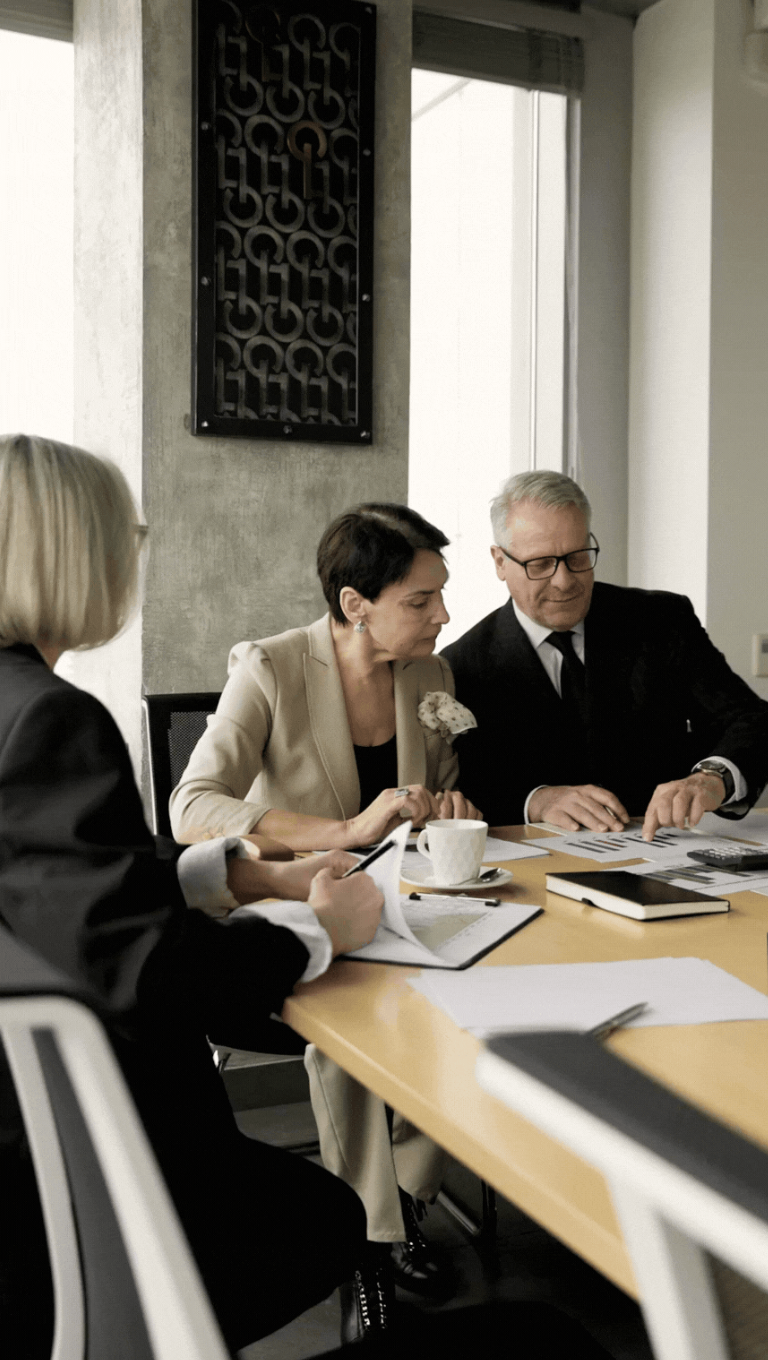Three people sitting at a conference table having a discussion, with documents, notebooks, and coffee cups in front of them.