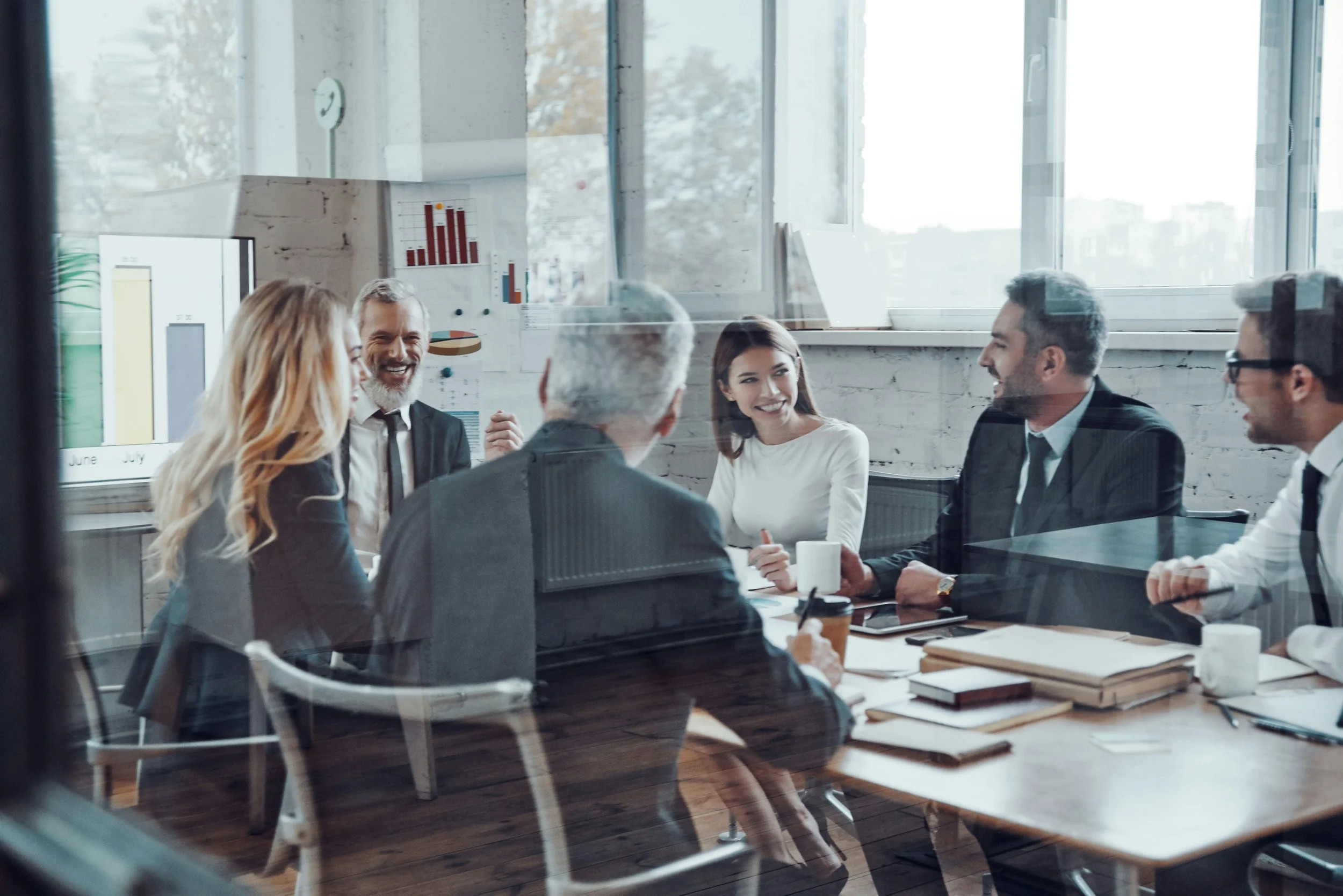 A group of seven diverse people sitting around a conference table, smiling and engaged in a discussion in an office with large windows and charts on the wall.