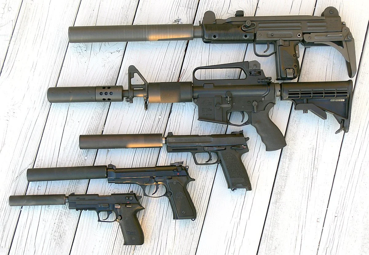 Four firearms including two handguns, a rifle with a silencer, and a submachine gun with a silencer, arranged on a light wooden floor.