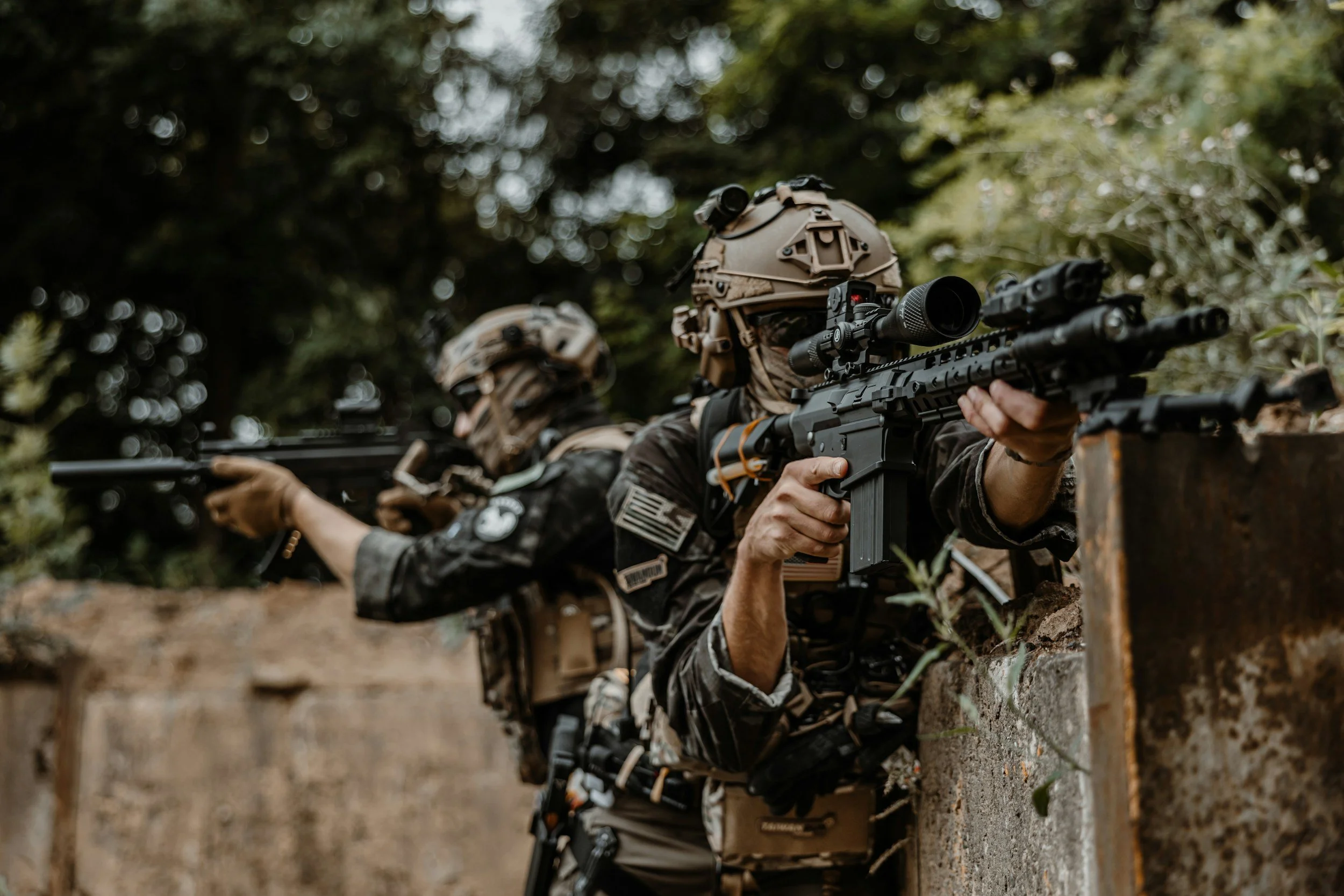 Two soldiers wearing camouflage uniforms and helmets aiming rifles while crouching behind a brick wall, in a wooded outdoor setting.