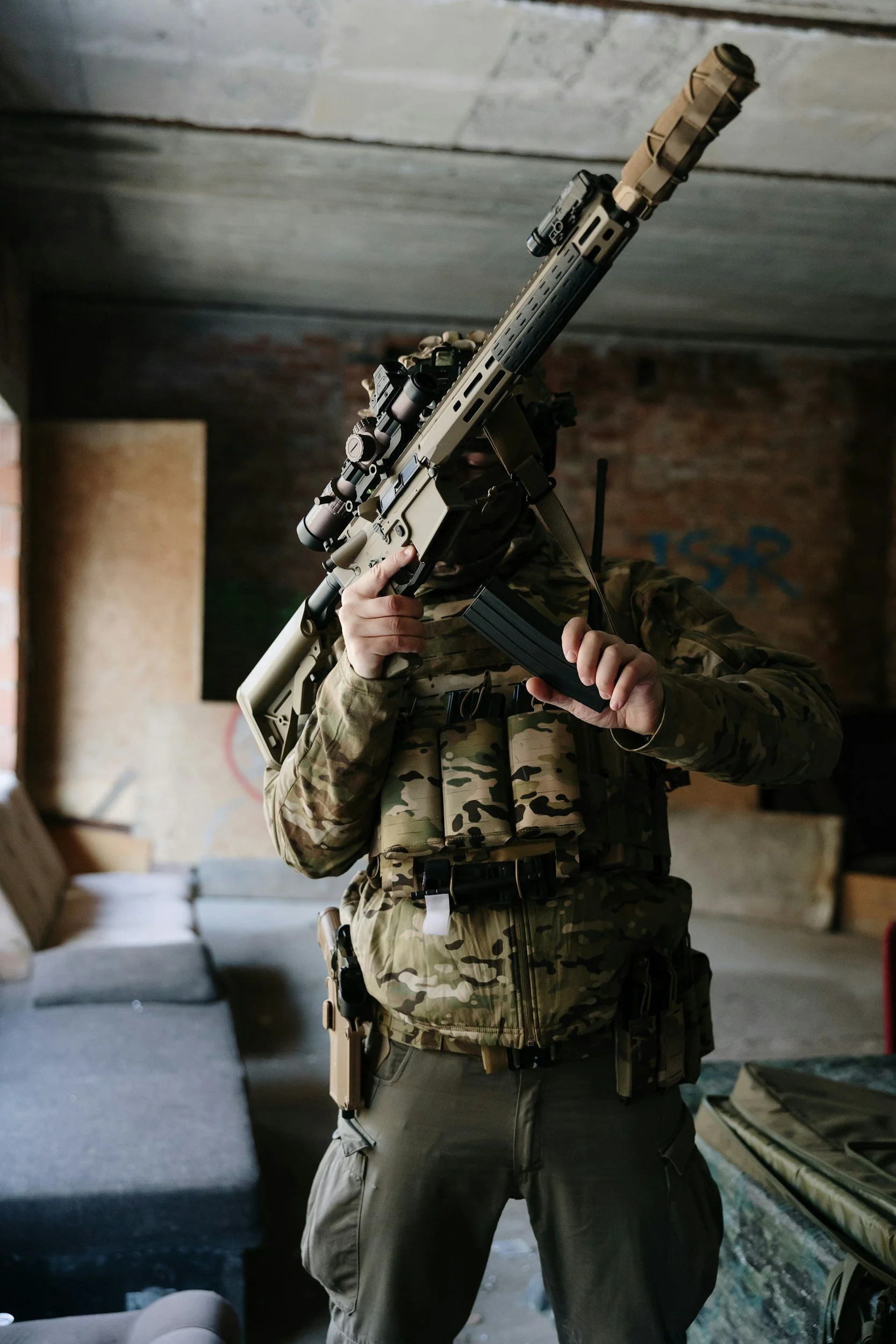 A person in camouflage military gear holding a sniper rifle, standing inside a room with concrete and brick walls.