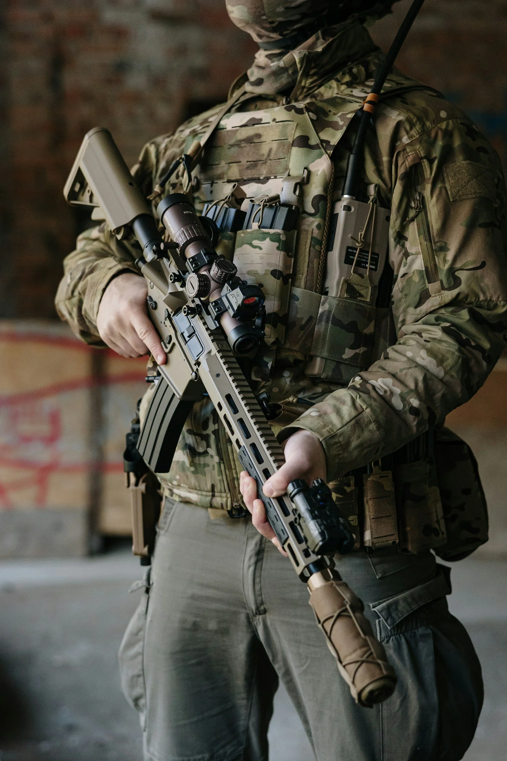 Soldier in camouflage uniform and tactical gear holding a rifle in an indoor setting.
