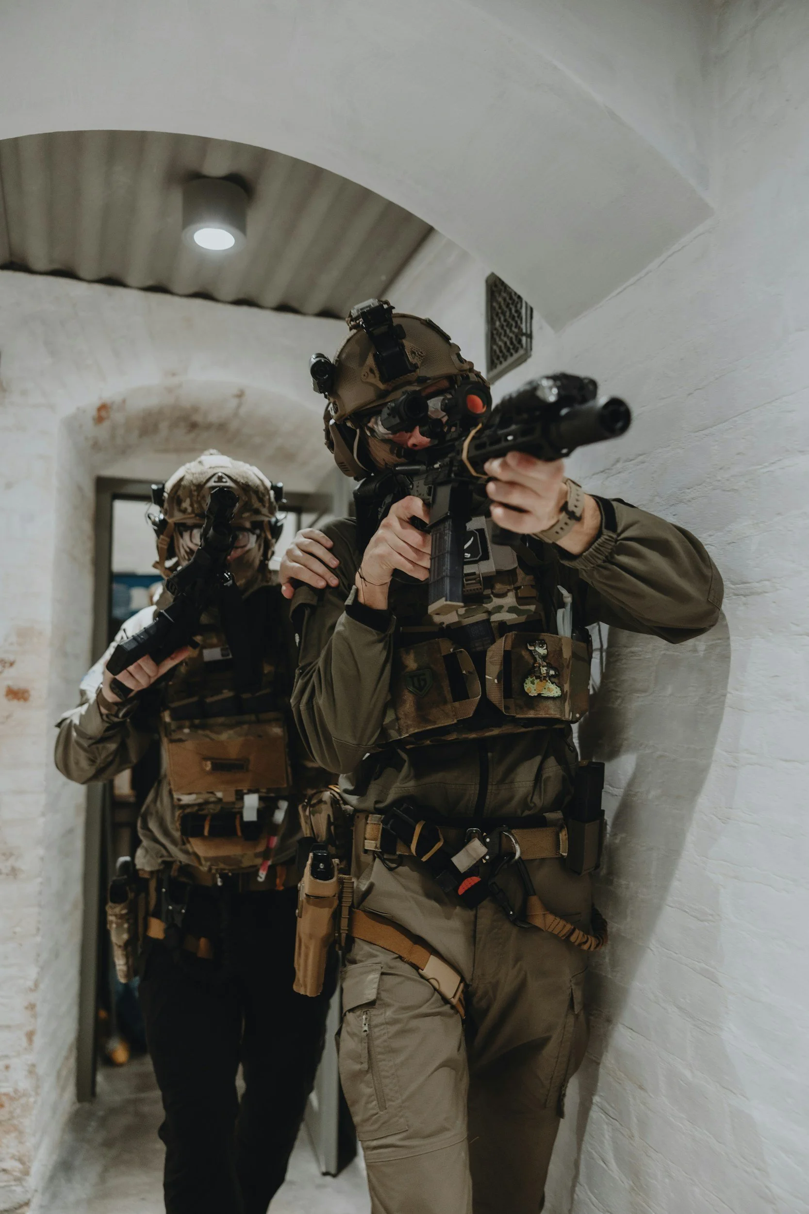 Two soldiers in tactical gear and helmets aiming guns down a corridor with white brick walls.