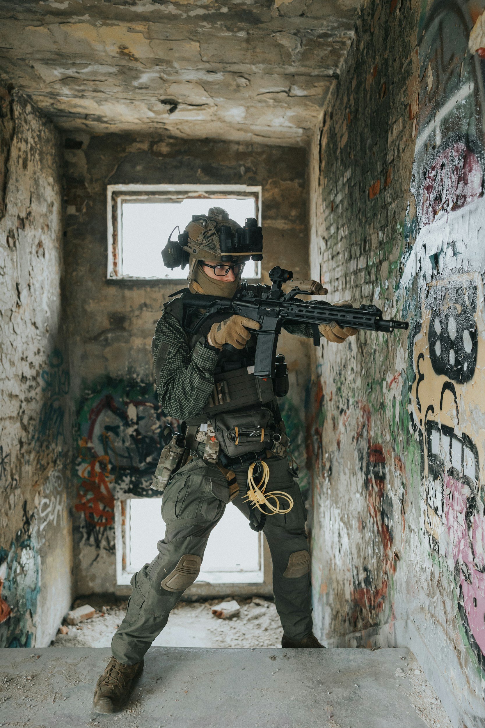 A person wearing military gear, including helmet, goggles, camouflage clothing, and gloves, aiming a rifle in an abandoned, graffiti-covered building with a large window and a damaged concrete floor.
