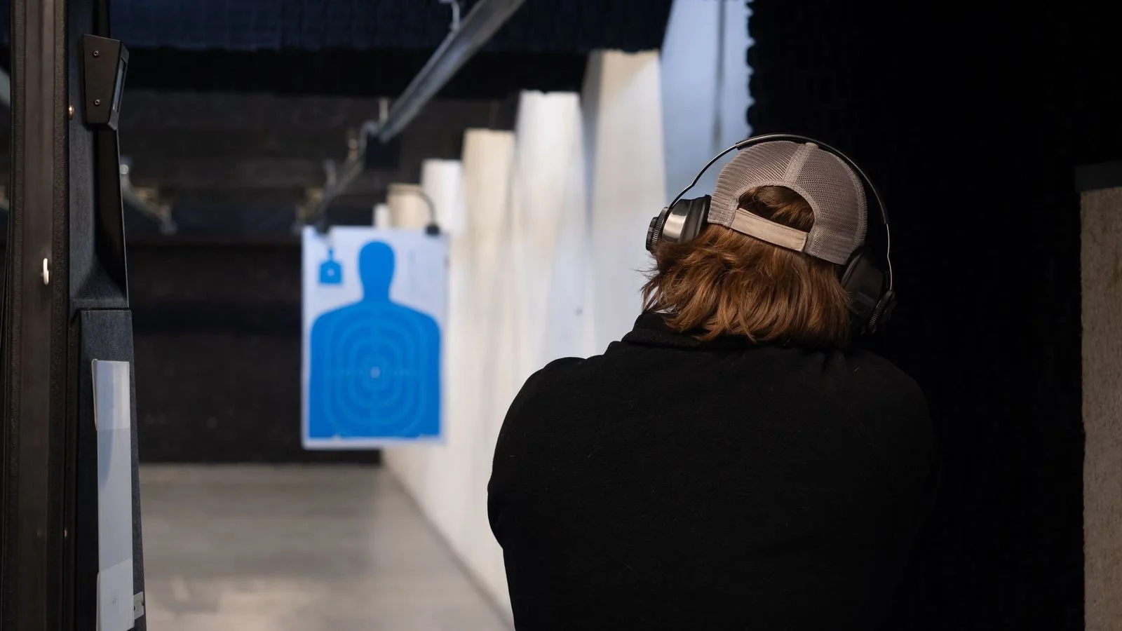 Person with long hair wearing a cap and headphones, aiming a firearm at a target in an indoor shooting range.