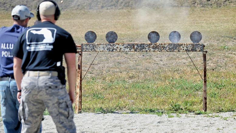 Two people at a shooting range observing the target with five metal disks on a rusted bar, some smoke rising from the right side.