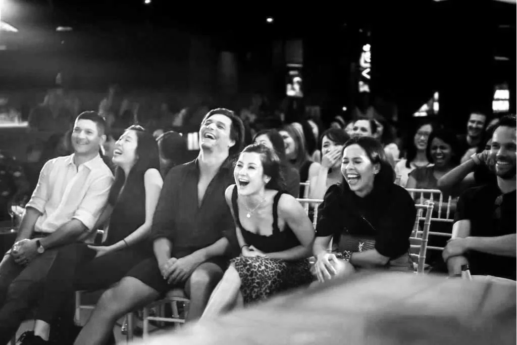 A group of people sitting in an audience, laughing and enjoying a performance or event at night.