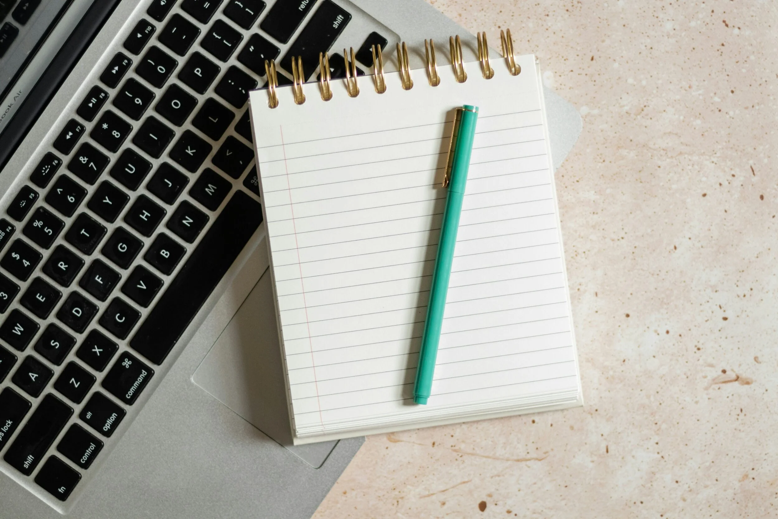 Close-up of a computer keyboard, a spiral notepad with lined paper, a teal pen, and a gold-trimmed pen on a beige speckled surface.