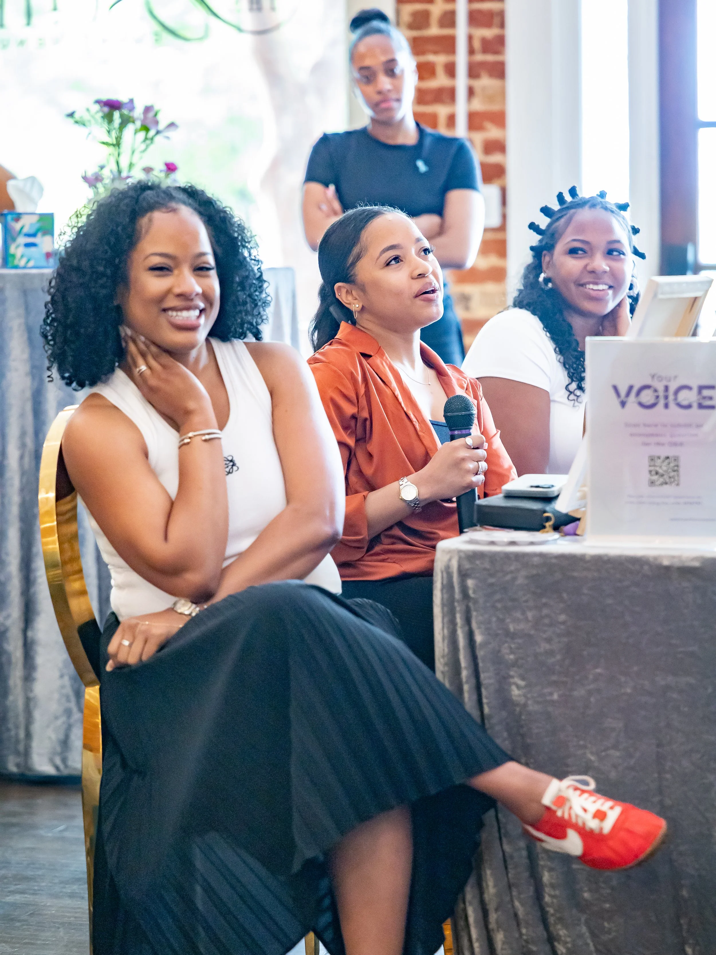 Four women attending a conference or meeting, one of whom is holding a microphone, with a third sitting on a yellow chair and wearing red sneakers.