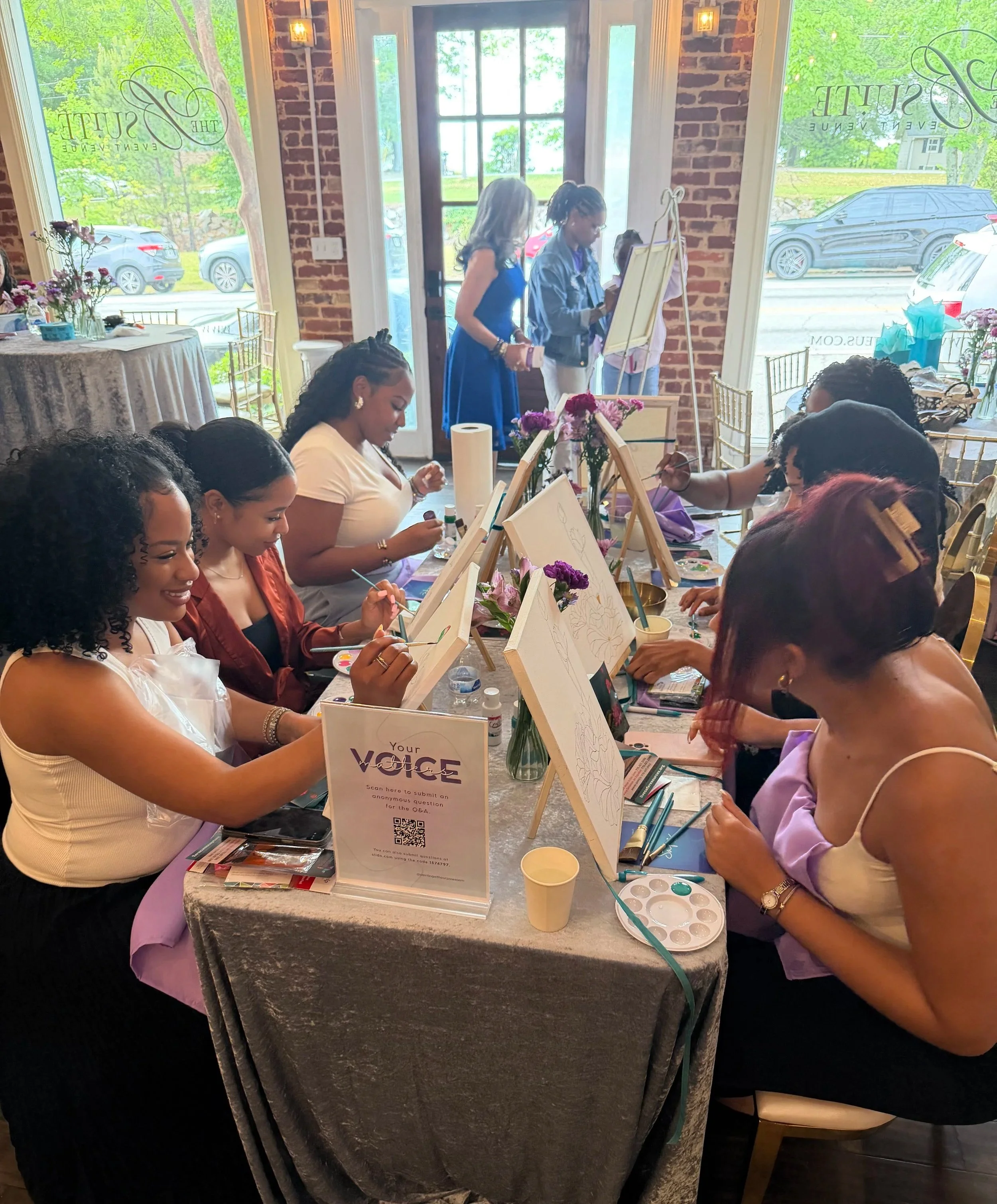 A group of women painting at a table during a craft event in a bright room with large windows, floral arrangements, and a brick wall. Some women are facing the table, focused on their canvases, while others are standing and chatting.