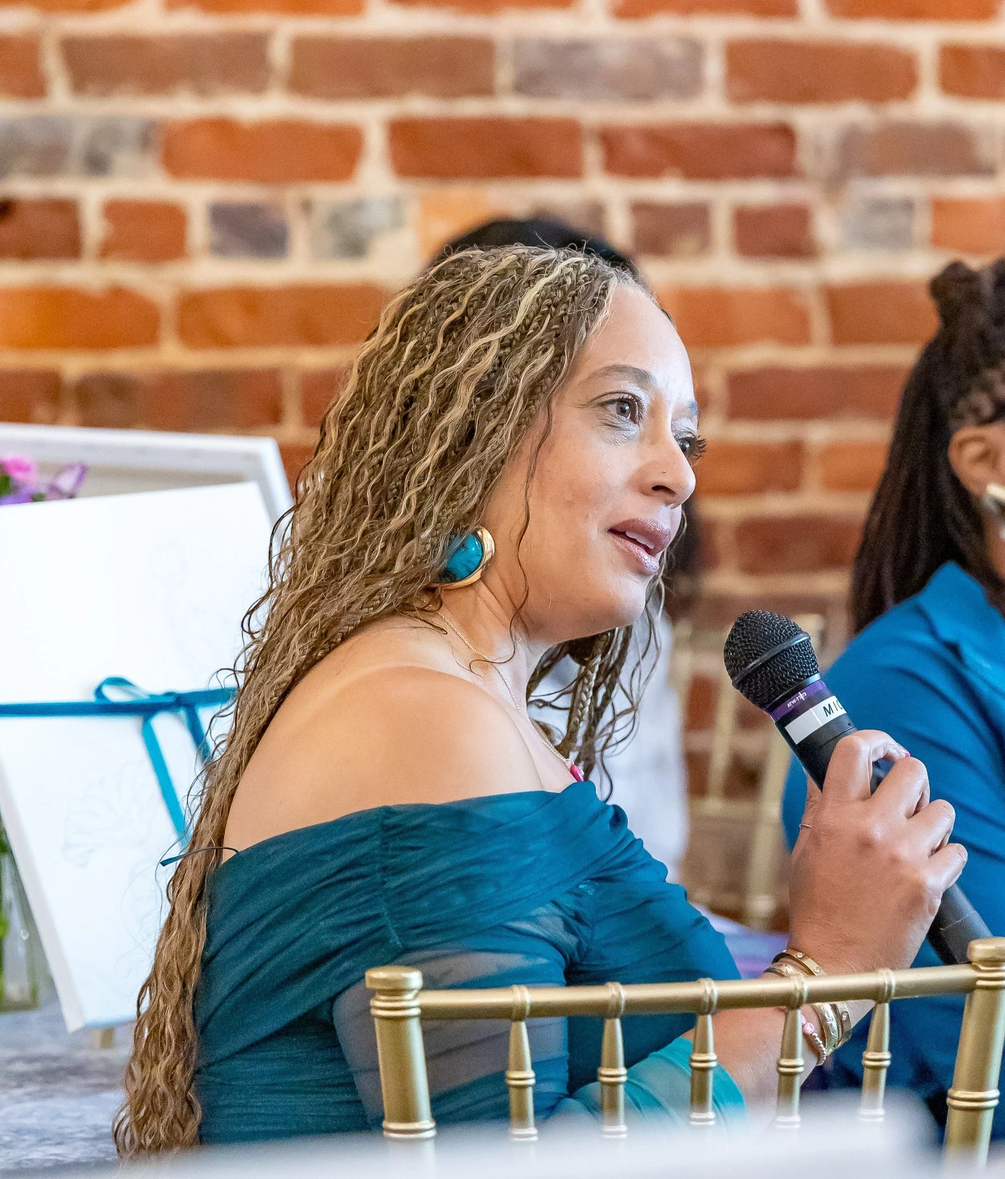 Woman with long, curly hair wearing large turquoise earrings and a blue off-shoulder dress, holding a microphone at a formal event with a red brick wall background.