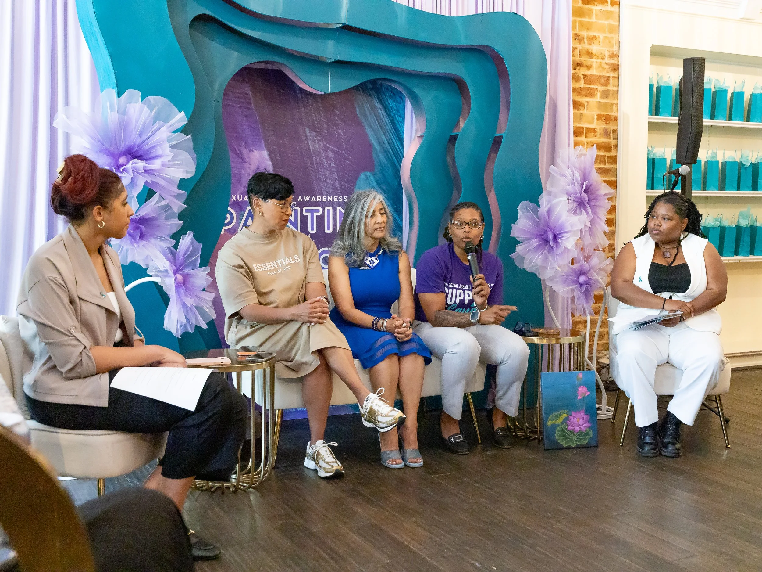 Five women sitting on chairs participating in a panel discussion or event, with a decorative backdrop and a brick wall behind them.