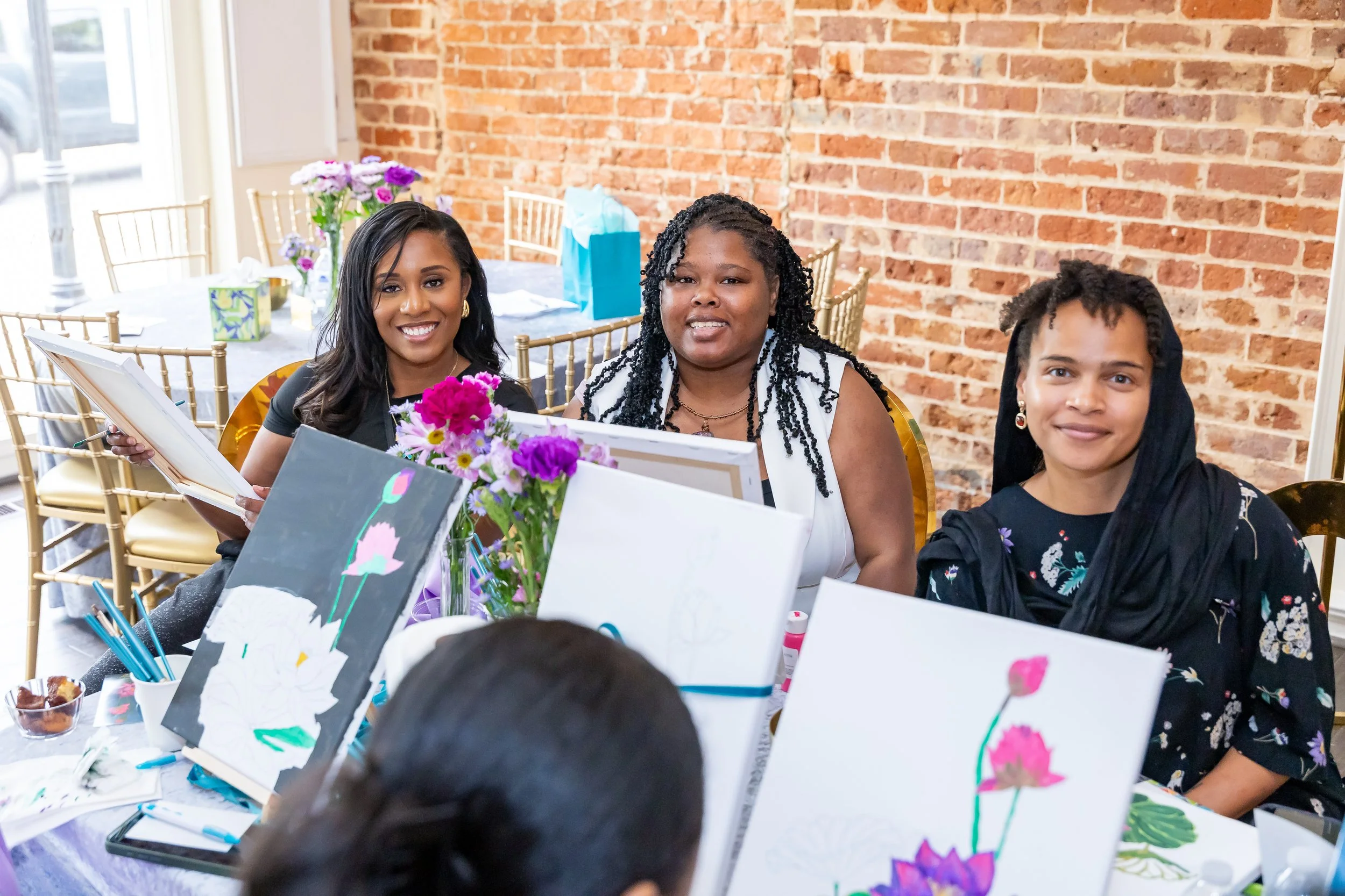 Three women sit at a table with artwork and flowers, smiling at an art event, with an exposed brick wall in the background.