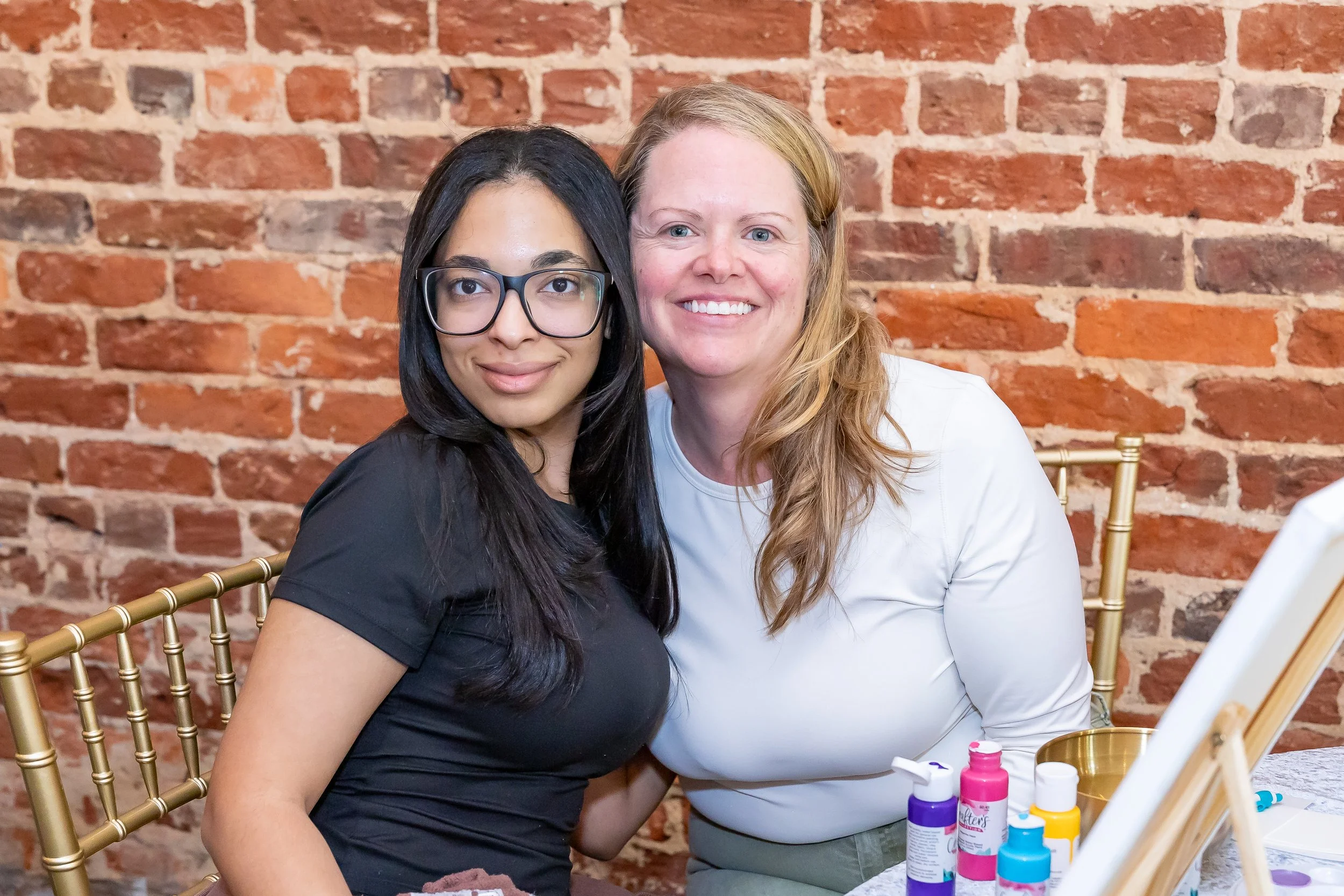 Two women sitting together at a table with art supplies, posing for a photo against a brick wall. One woman is wearing glasses and a black shirt, the other has curly blonde hair and is wearing a white shirt. There are bottles of paint and brushes on the table.