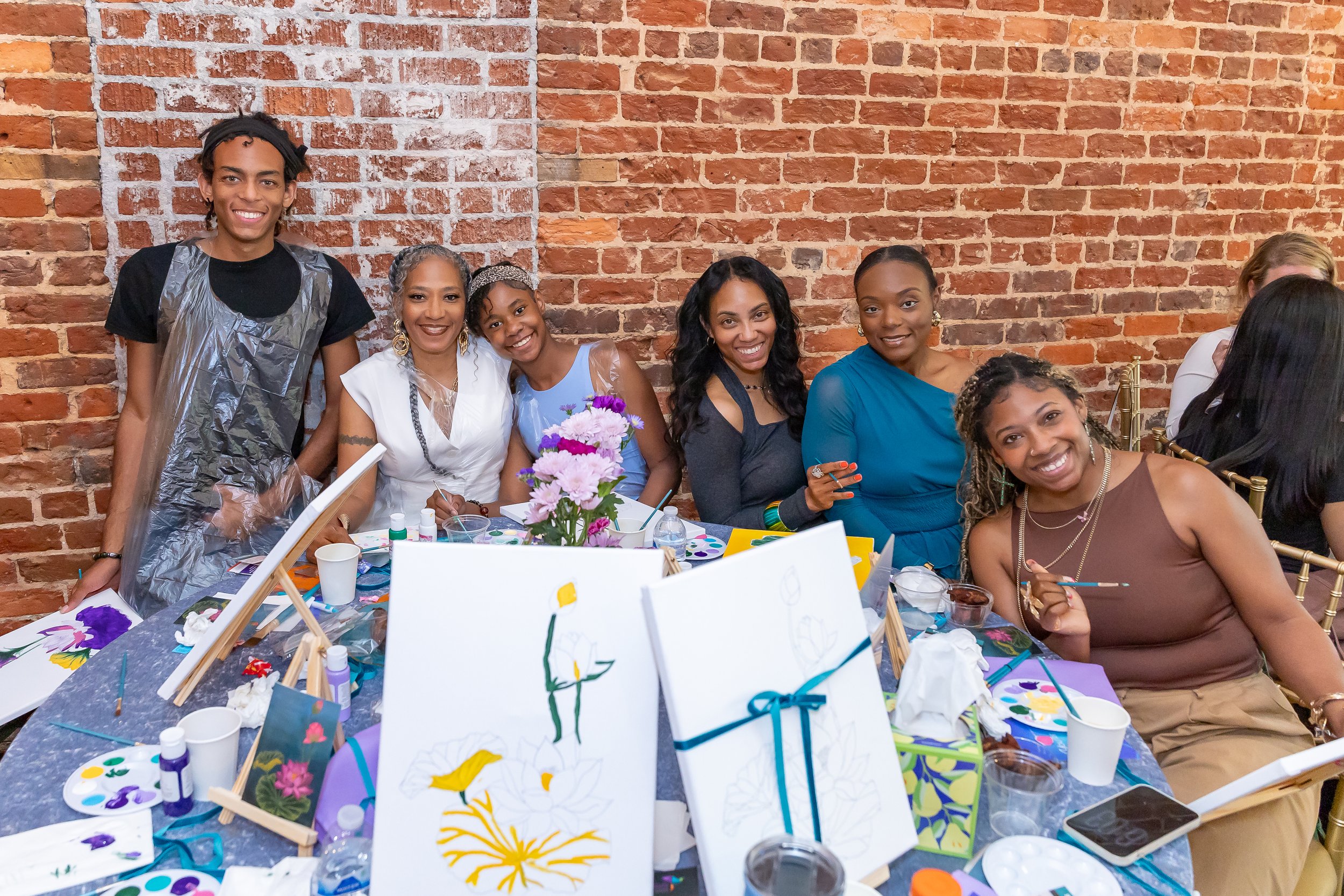 Group of women and a young man sitting at a table in an art workshop or party, with art supplies, paintings, and a flower centerpiece against a brick wall background.