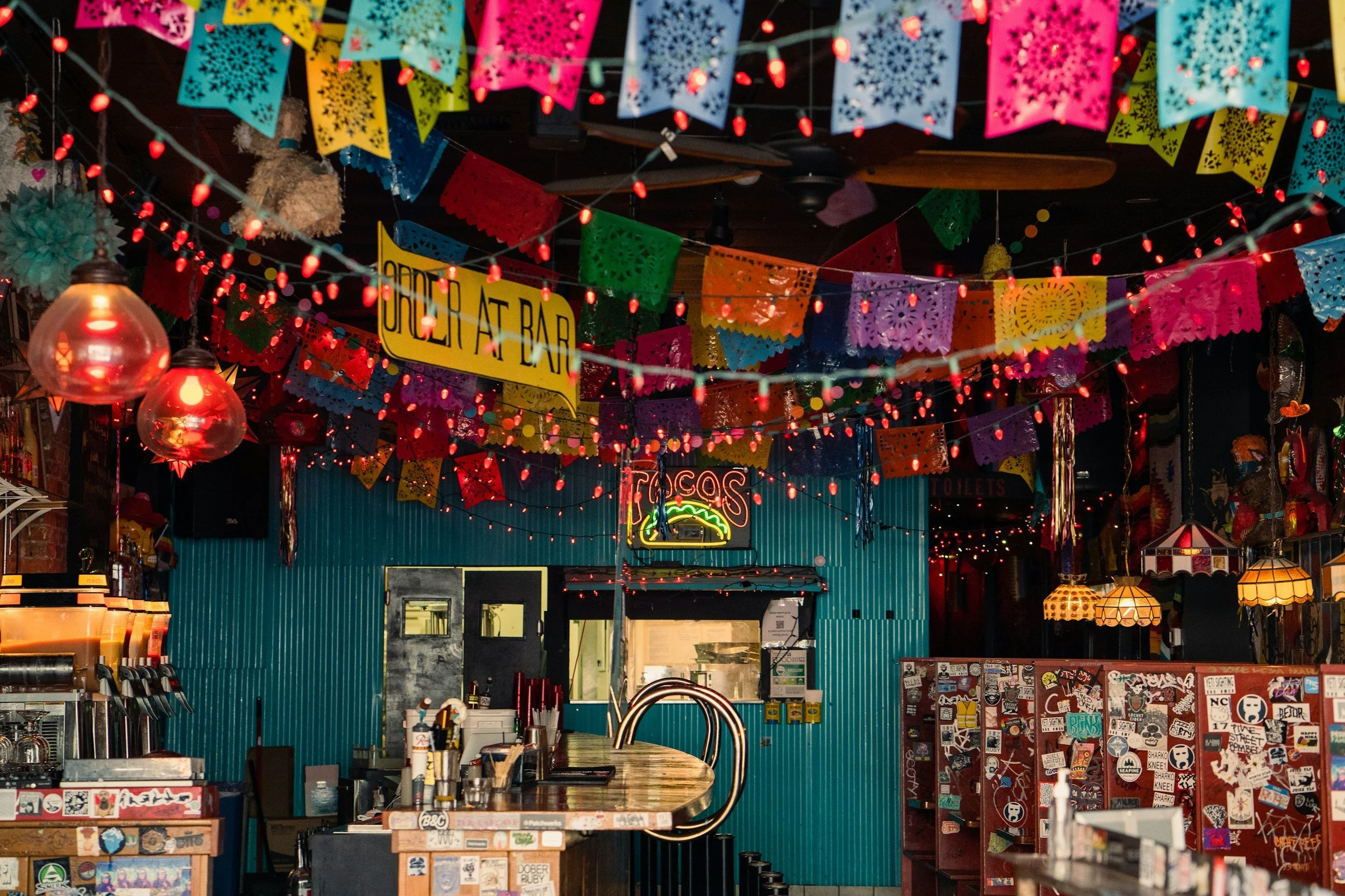 Colorful papel picado banners and string lights hanging above a bar area, with a neon taco sign in the background in a festive Mexican restaurant or bar.