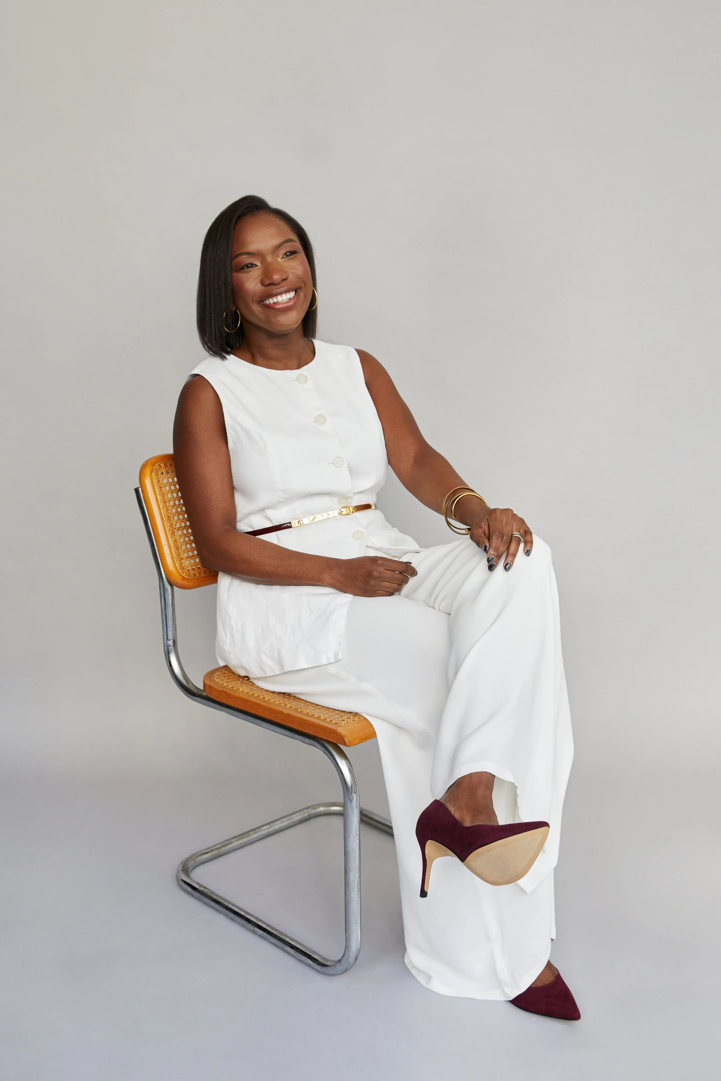 A woman sitting on a chair with a light grey background. She has medium-length black hair, wearing a white sleeveless dress with gold buttons and a brown belt. She has hoop earrings, bracelets, and rings, and is smiling while looking slightly to her right.