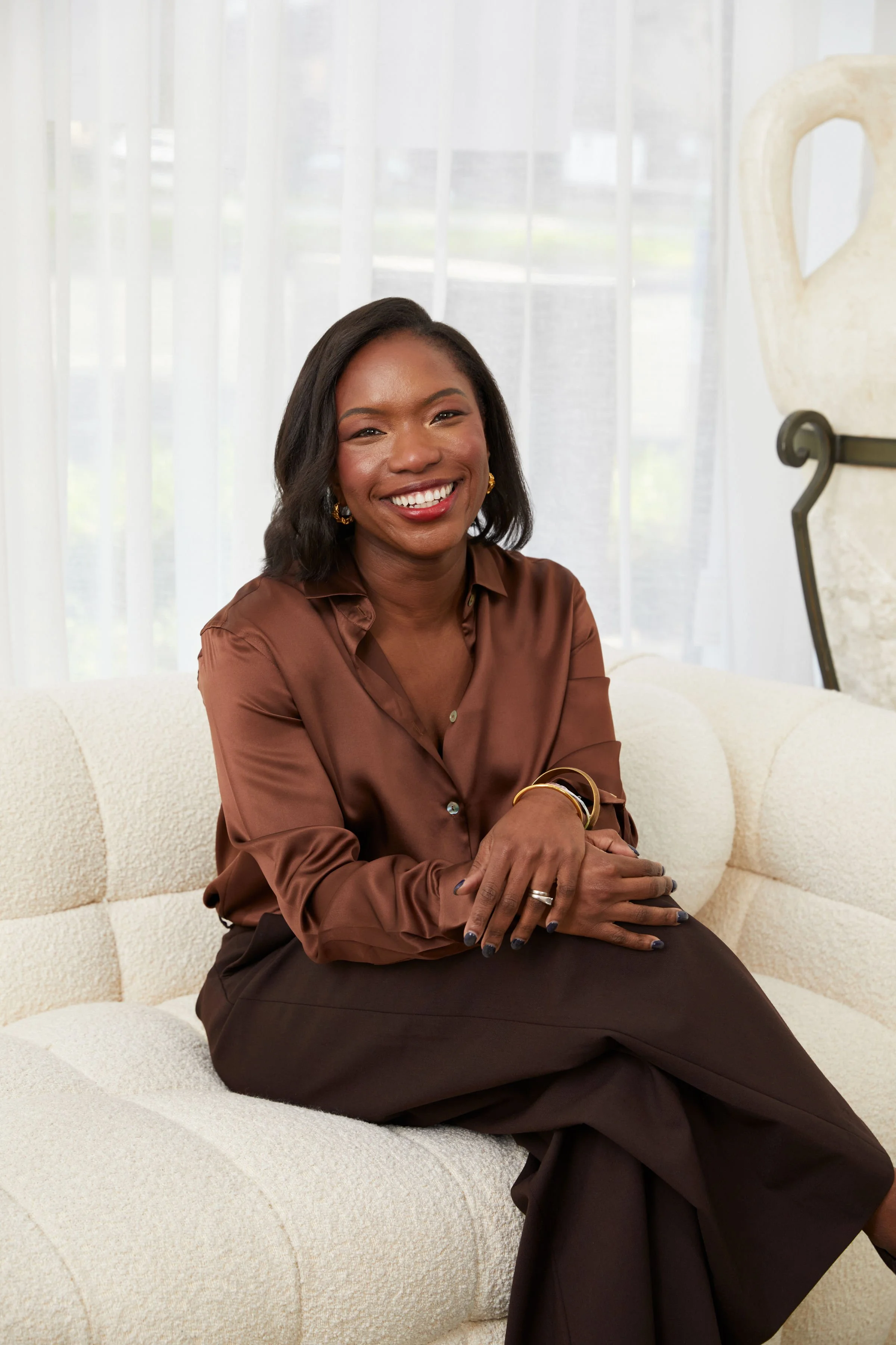 A woman with dark hair wearing a brown blouse and brown pants, seated on a cream-colored sofa in a room with white curtains and a decorative sculpture in the background, smiling at the camera.