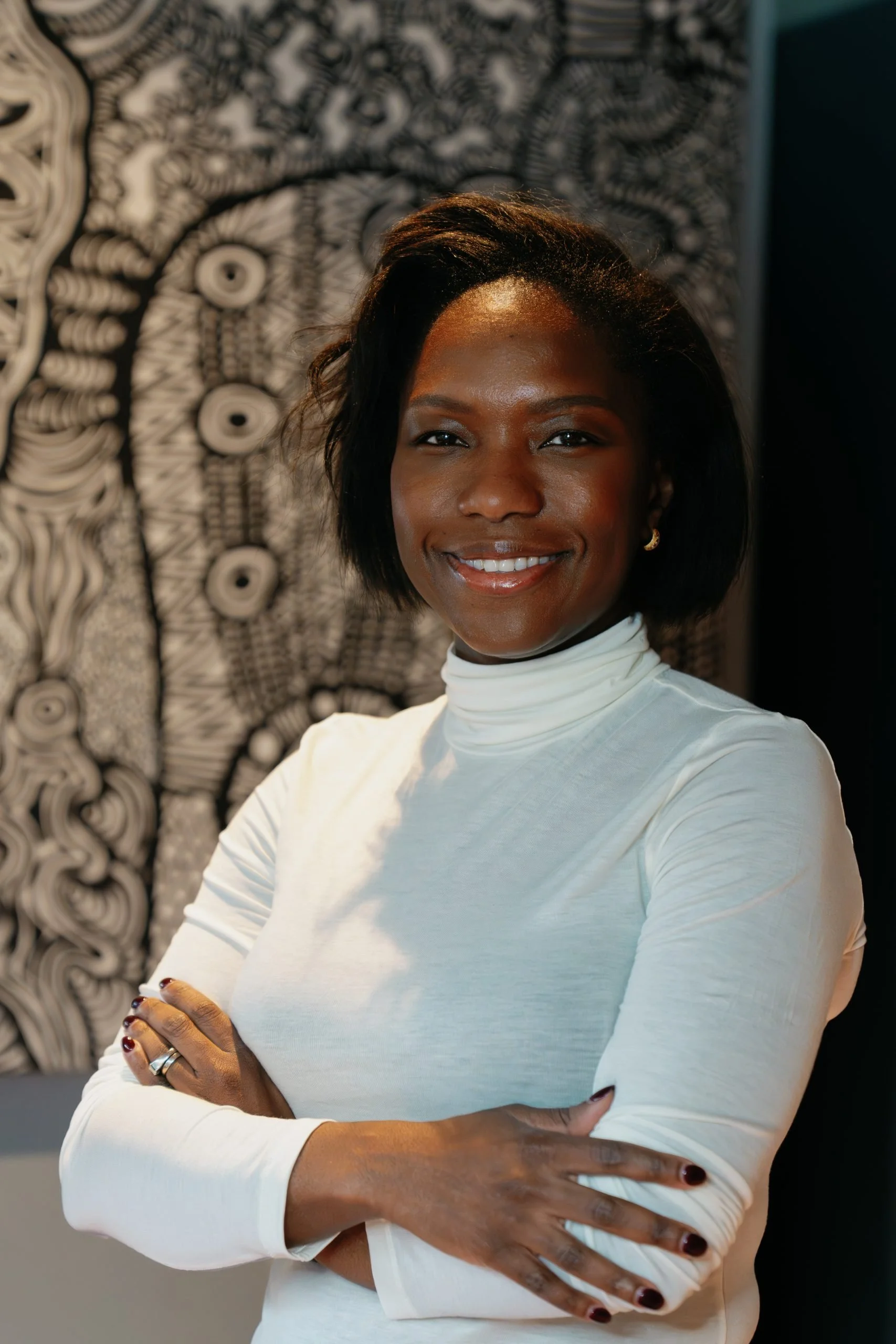 A woman with dark hair and a wide smile wearing a white turtleneck shirt, standing with her arms crossed in front of a black and white abstract art background.