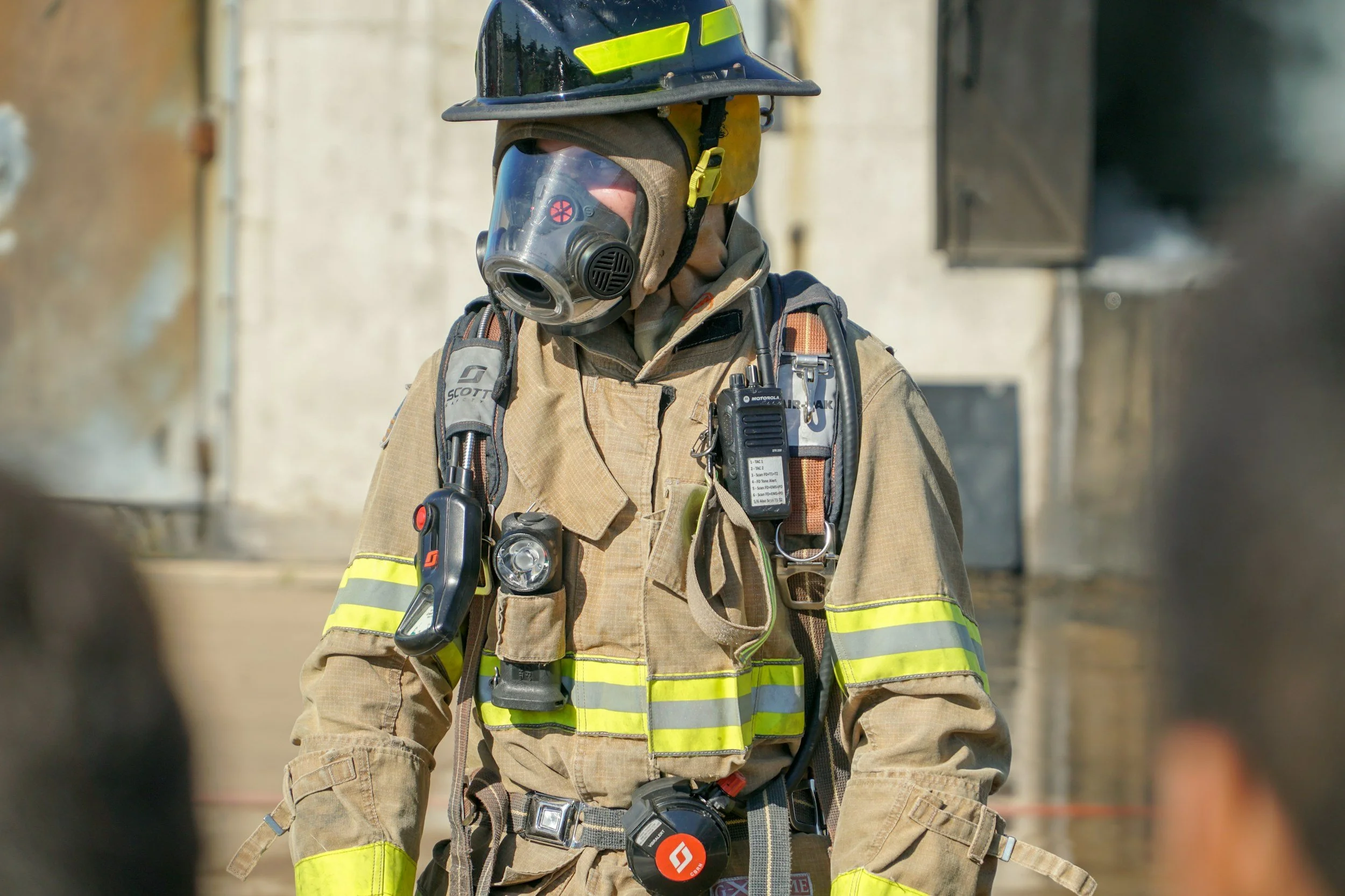 A firefighter dressed in tan turnout gear with yellow reflective stripes, wearing a black helmet with yellow markings, a protective mask, and holding equipment, standing outdoors with a blurred urban background.