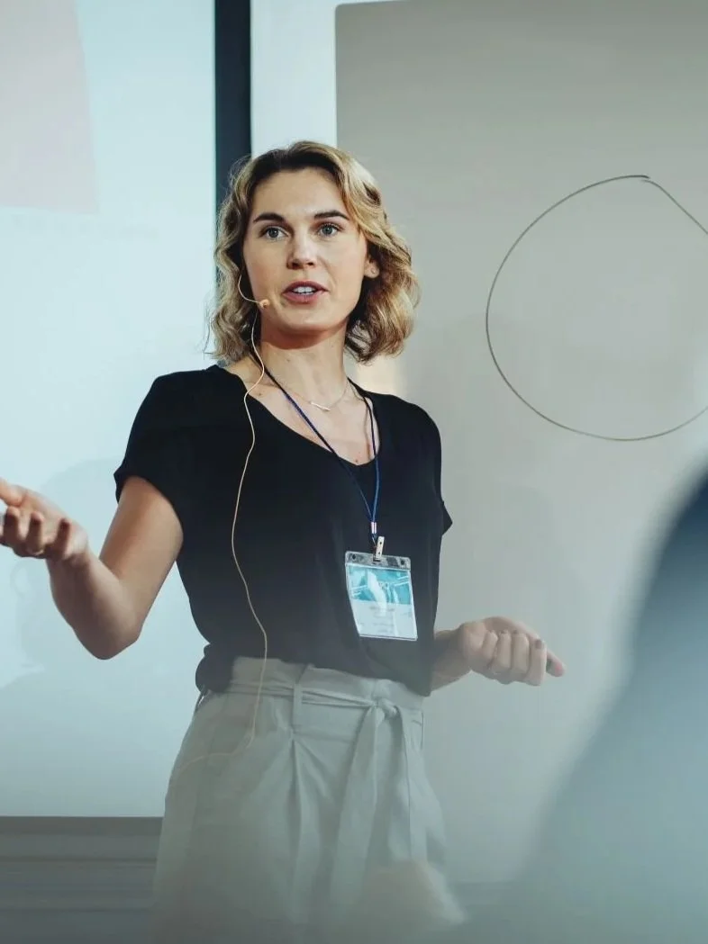 A woman with shoulder-length wavy blonde hair giving a presentation in front of a whiteboard, wearing a black top, beige pants, and a conference badge around her neck.
