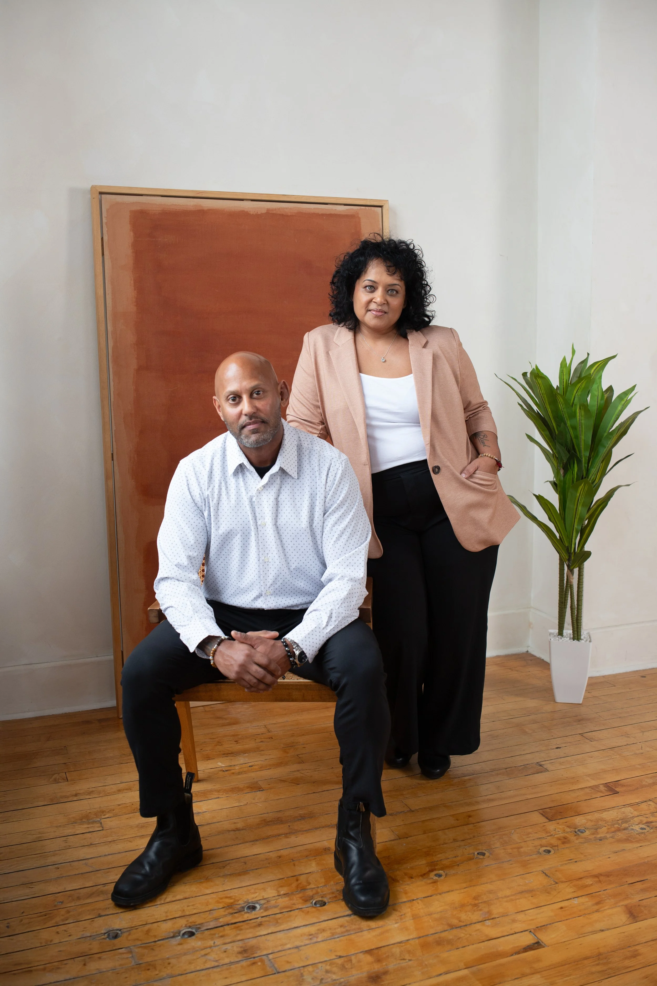 A man with a shaved head and beard wearing a white patterned shirt and black pants is sitting on a wooden chair with his hands clasped. A woman with curly black hair, wearing a tan blazer, white shirt, and black pants, stands beside him with one hand on her hip. They are in a room with wooden flooring, a brown abstract painting behind them, and a tall green potted plant to the right.