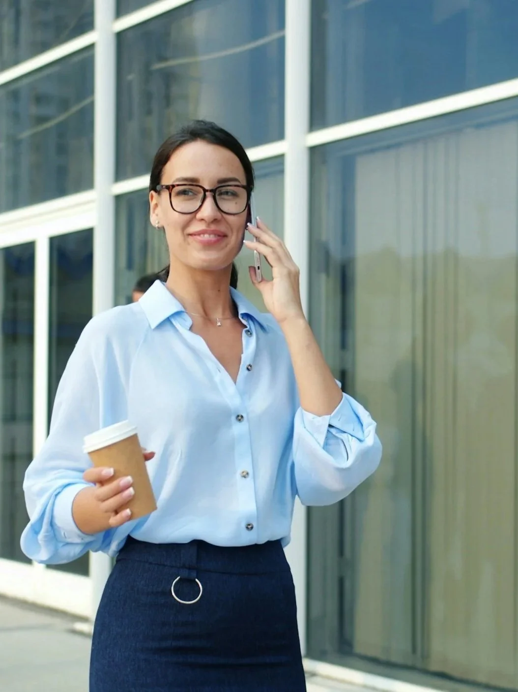 A smiling woman with glasses, wearing a blue button-up shirt and dark skirt, holding a coffee cup in one hand and talking on her cellphone outside a modern office building.