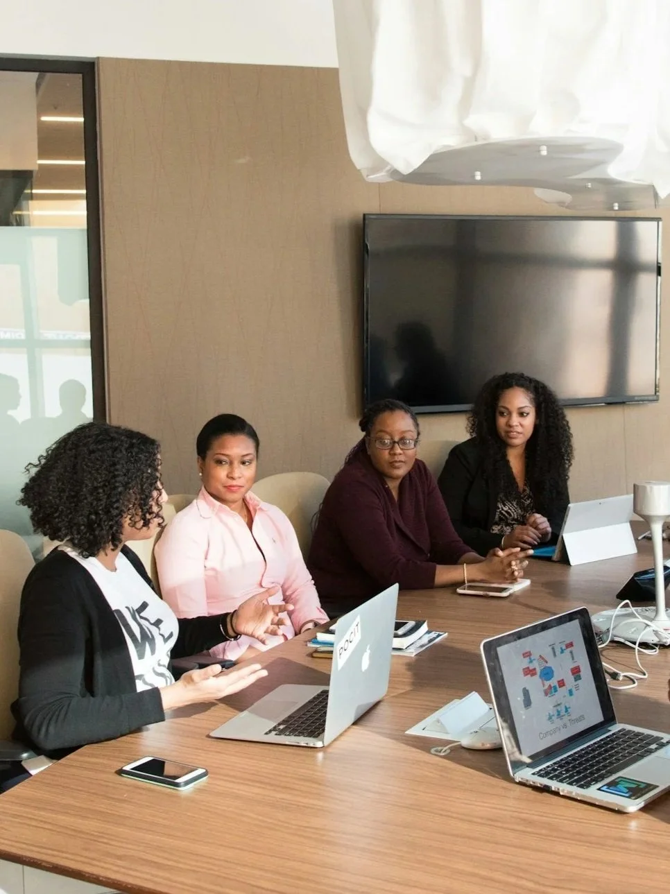 Four women sitting at a conference table during a meeting, with laptops, notebooks, and a large screen on the wall
