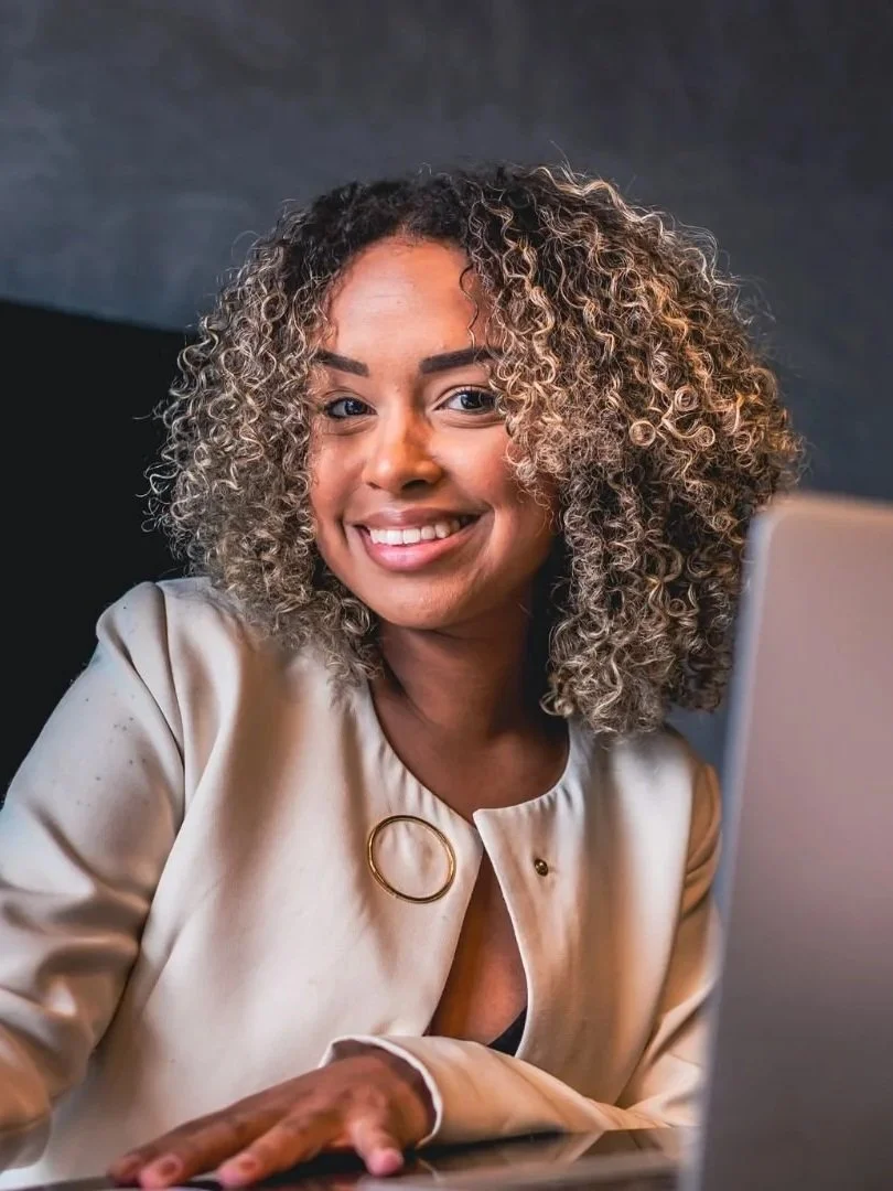 A smiling woman with curly hair using a laptop.