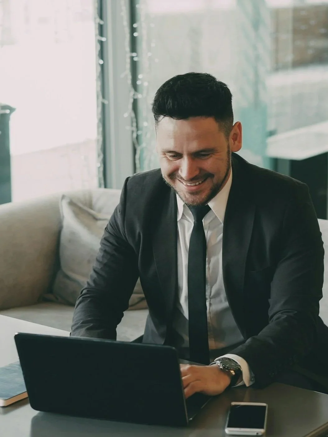 A man in a black suit and tie sitting at a table, smiling while looking at a laptop in front of him.