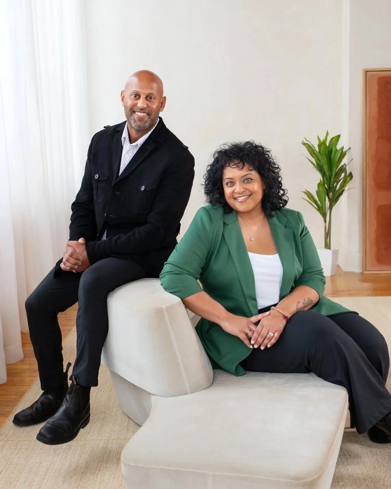 A man in a black jacket and a woman in a green blazer sitting on a modern beige couch in a well-lit room with a potted plant and wooden cabinet.