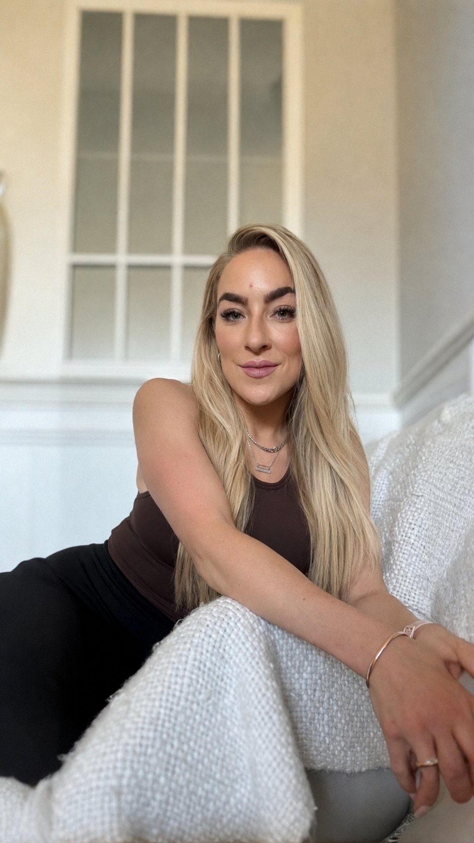 A young woman with long blonde hair, wearing a black sleeveless top, is sitting on a white textured sofa in a well-lit room with a white wall and window in the background.