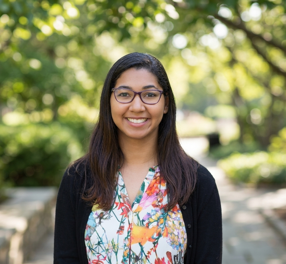 A young woman with glasses smiling outdoors on a sunny day, wearing a colorful floral top and black cardigan, with blurred greenery in the background.