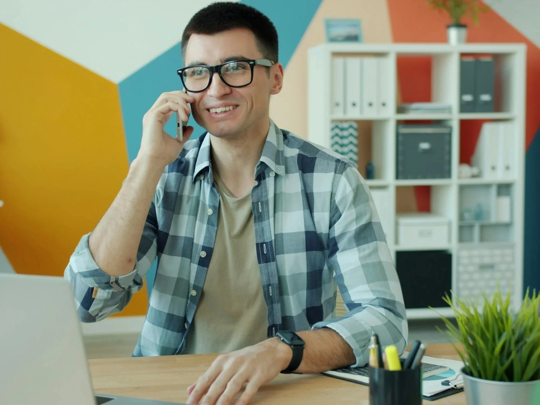 Young man with glasses smiling and talking on a cellphone in a modern office, wearing a plaid shirt and smartwatch.