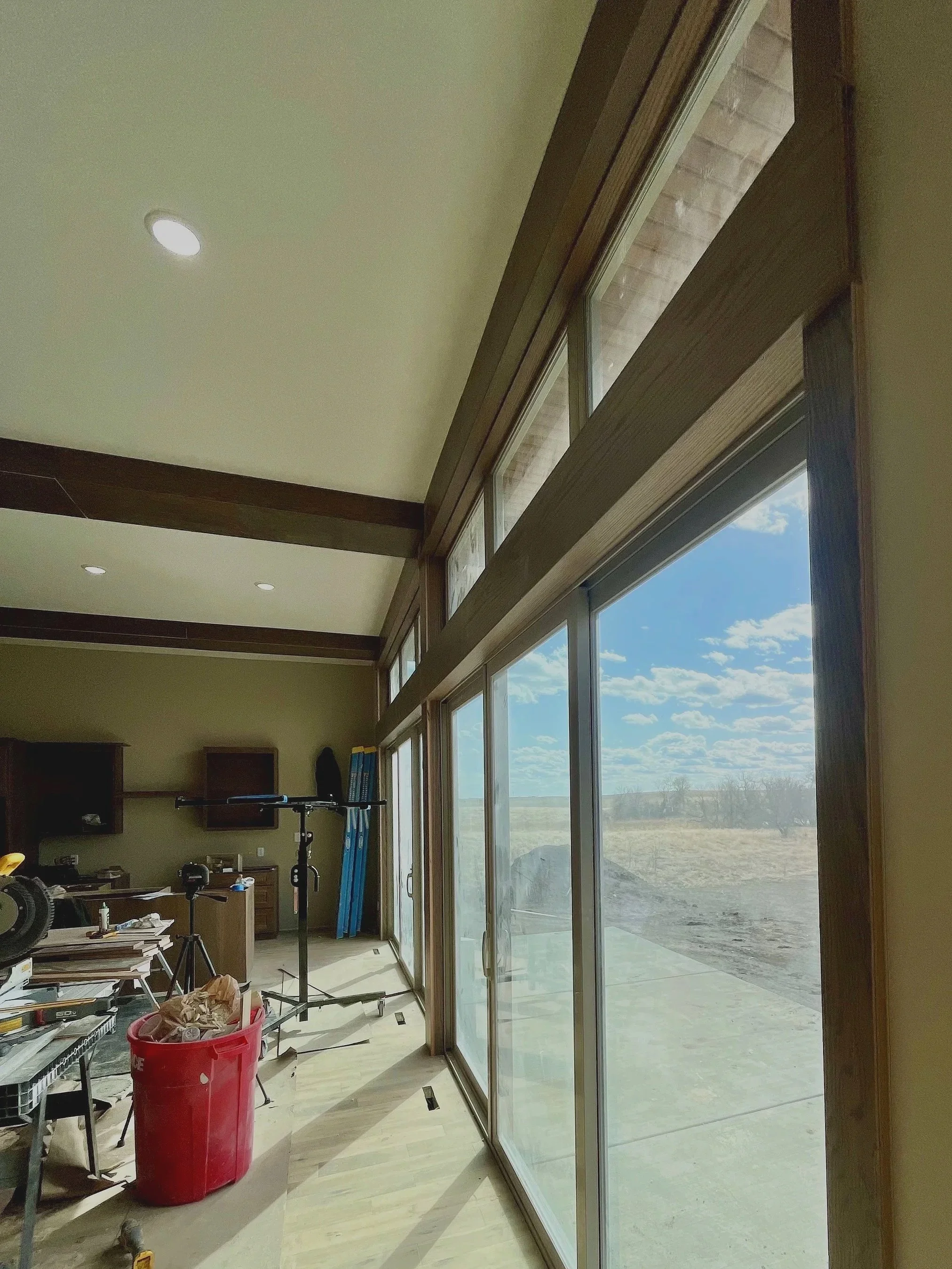 Interior view of a room with large sliding glass doors showing a outdoor landscape, with various tools, equipment, and a red trash bin inside.