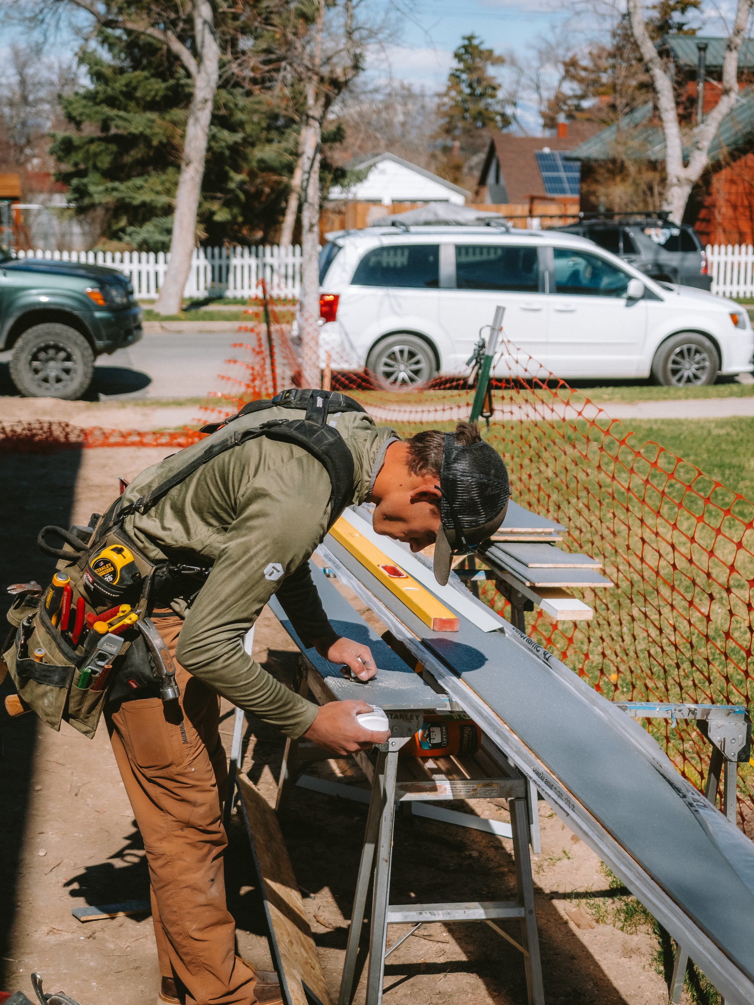 A man working on a solar panel, wearing construction tools and gear, outdoors with fencing, trees, and parked cars in the background.