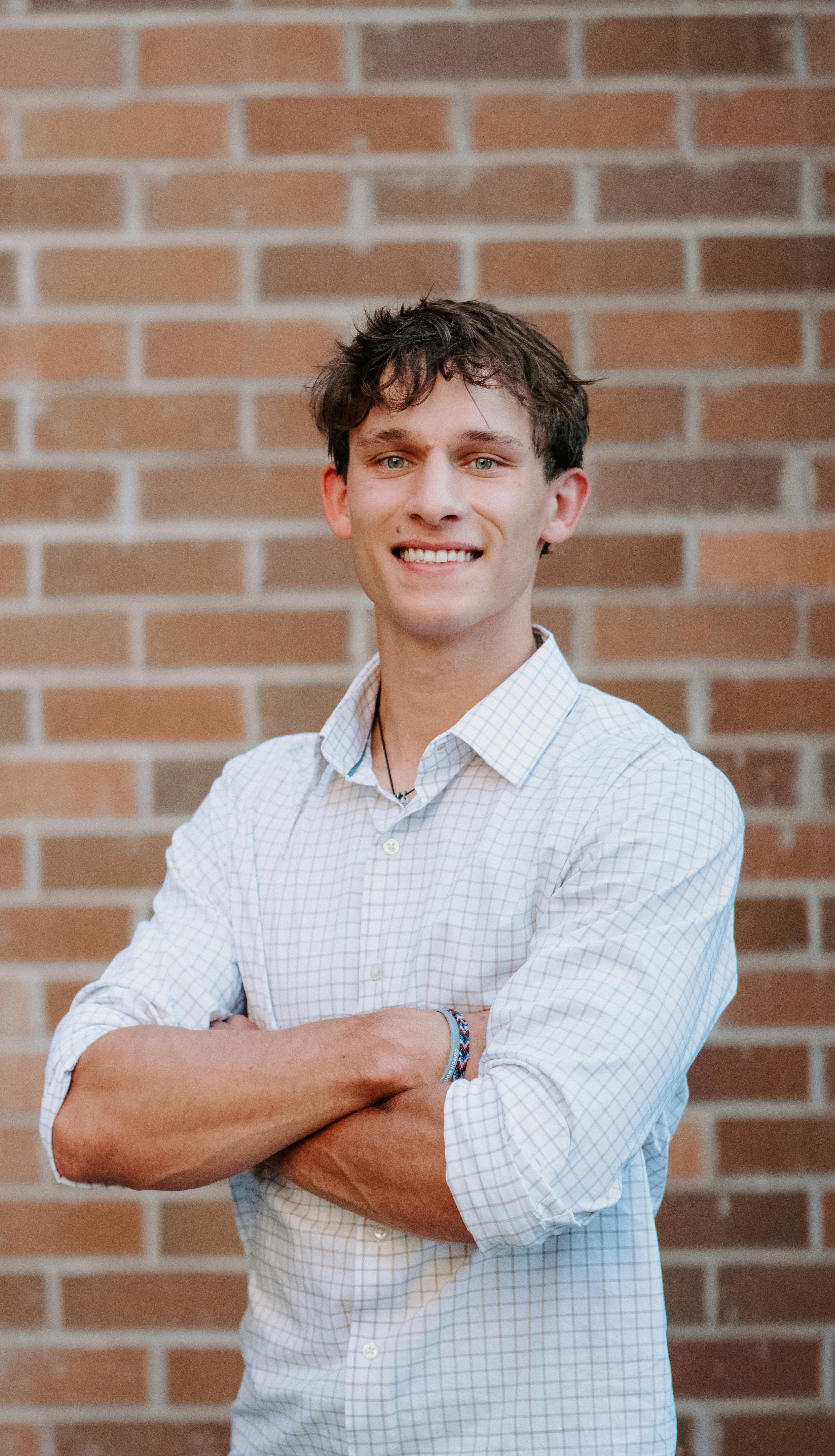 A young man with dark, tousled hair, wearing a white checkered shirt, standing in front of a brick wall, smiling with his arms crossed.