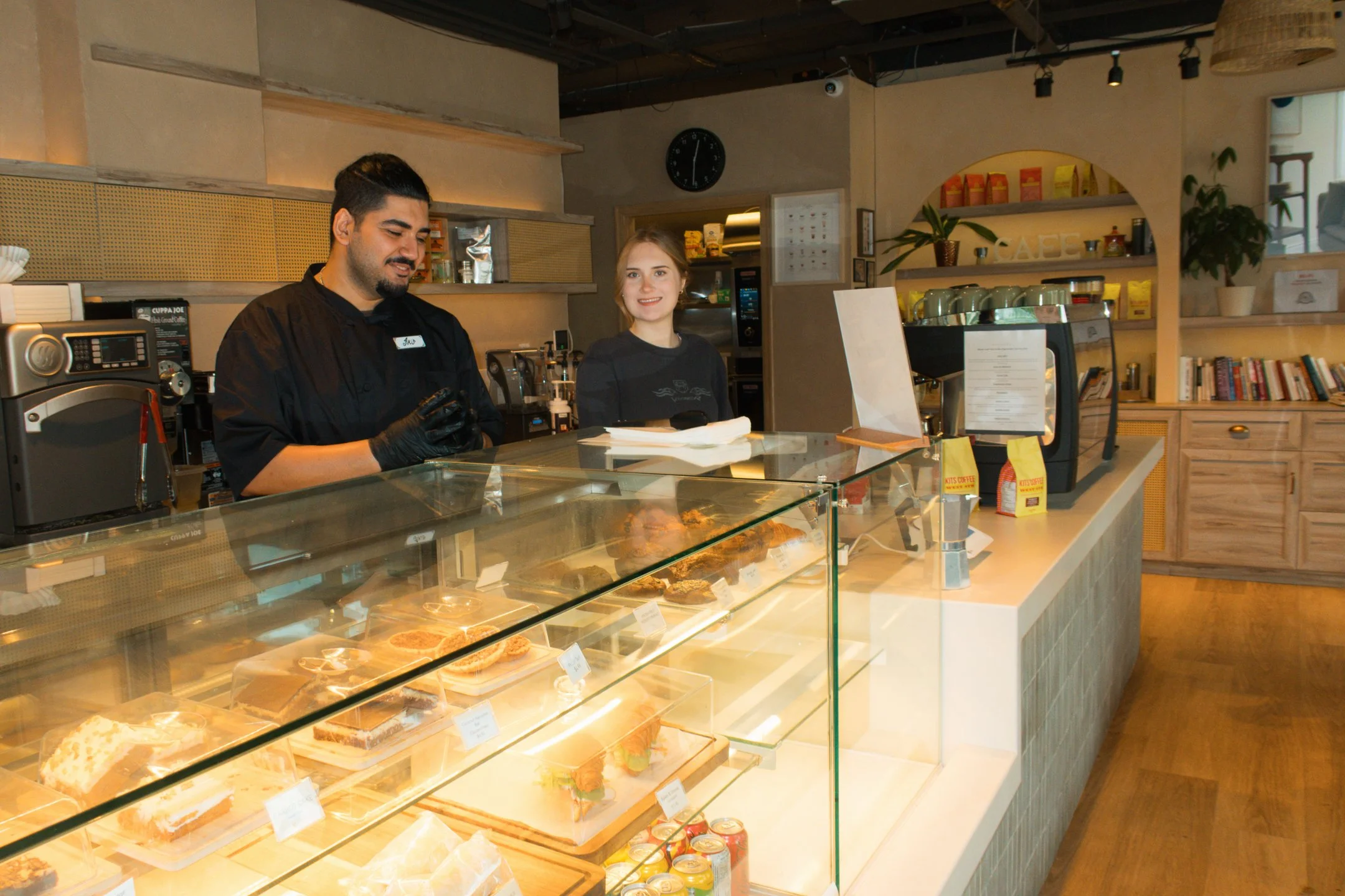 A man and a woman behind a display counter at a cafe, with baked goods in the glass case and shelves with books and plants in the background.