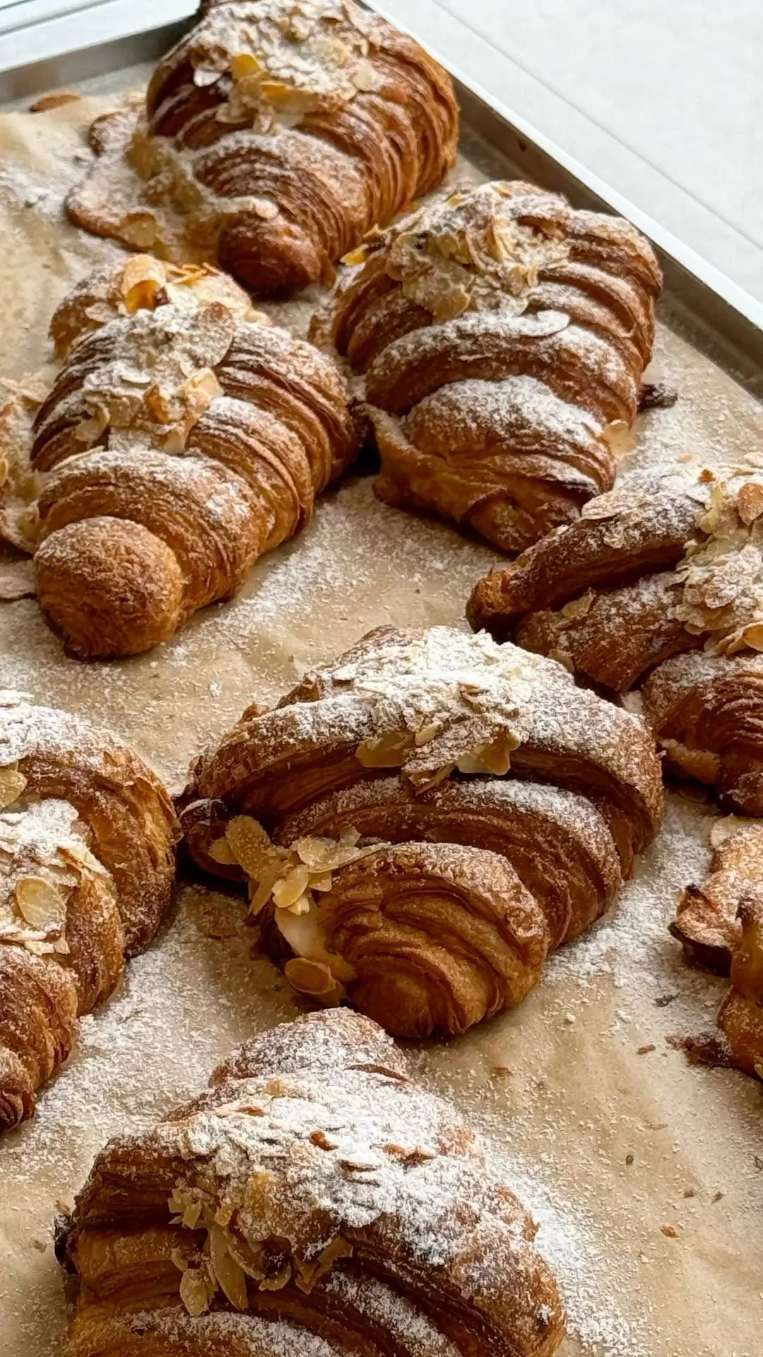Assorted croissants topped with powdered sugar and sliced almonds on a baking sheet.