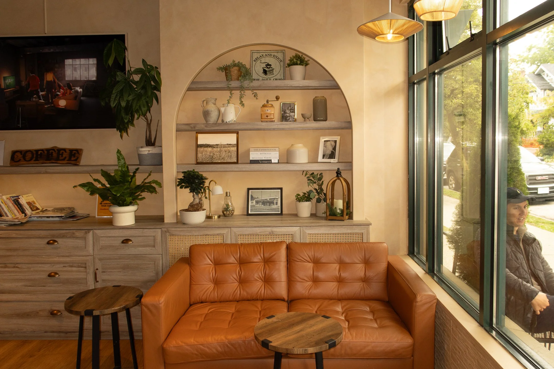 Interior of a cozy cafe or lounge with a tan leather sofa, small wooden tables, a light wooden cabinet with various decorative objects, books, and framed pictures, large windows showing a green outdoor scene, and hanging pendant lights.