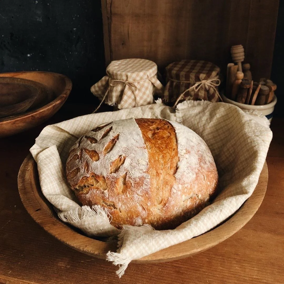 A round loaf of rustic bread with a dusting of flour on top, placed in a wooden bowl lined with a cloth, with jars and wooden utensils in the background.