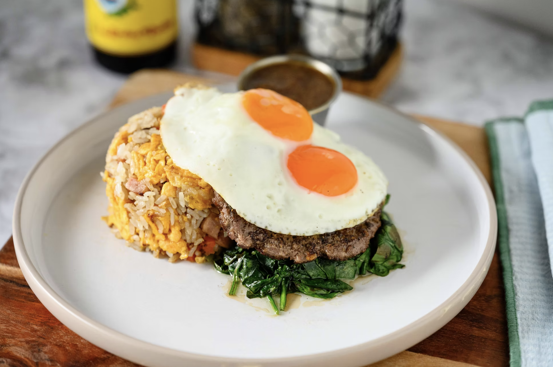 A plate with fried rice topped with two fried eggs, a beef patty, and spinach leaves. In the background, there is a small cup of sauce, a bottle, and a metal wire basket on a wooden tray.