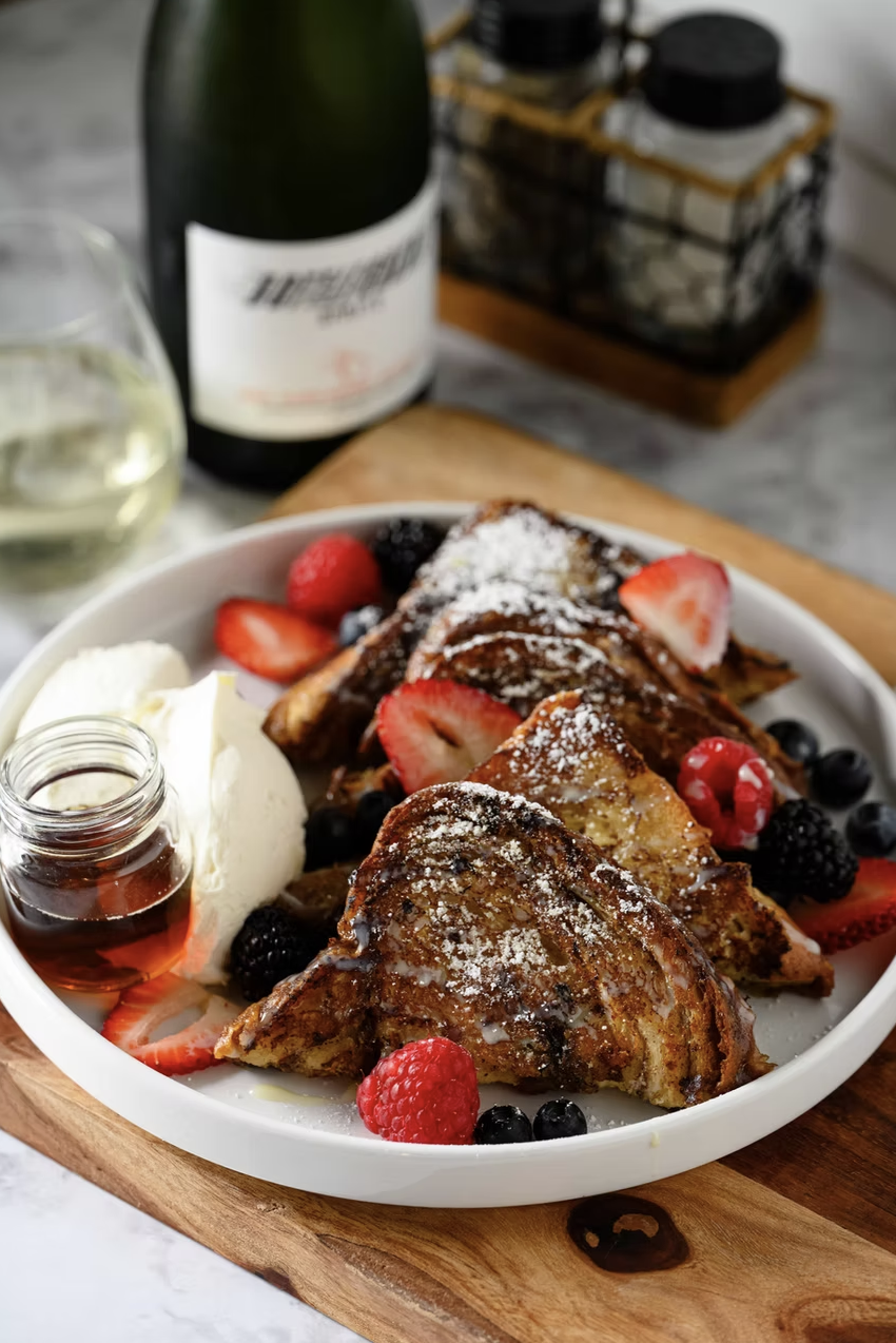A plate of French toast topped with powdered sugar and fresh berries, served with a side of whipped cream, syrup, and a glass of white wine.