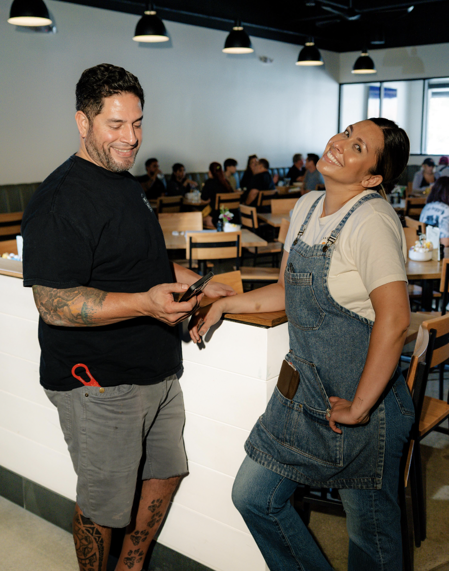 A man and a woman in a casual restaurant, smiling and chatting. The man is holding a phone and has tattoos on his arm and leg. The woman is wearing a white t-shirt and denim apron, leaning against a counter.