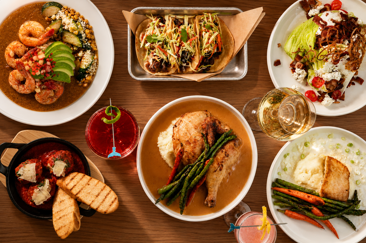 A table filled with various dishes including shrimp and avocado on a plate, tacos with shredded cheese and vegetables, a salad with lettuce, tomatoes, blue cheese, and bacon bits, a bowl of grilled chicken with mashed potatoes and green beans, a skillet of meatballs with bread slices, and other plated foods. There are also two glasses of white wine and a red cocktail.