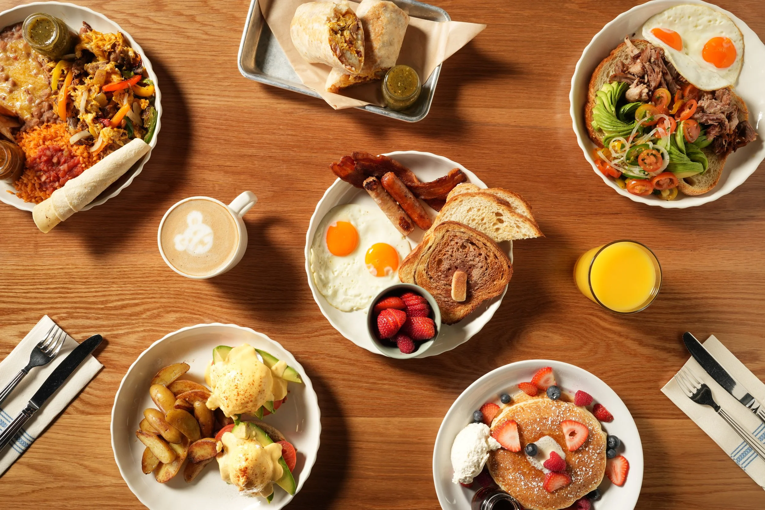 A variety of breakfast dishes on a wooden table including fried eggs, pancakes with berries, breakfast burrito, avocado toast, potatoes, sausage, bacon, strawberries, blueberries, orange juice, and coffee.
